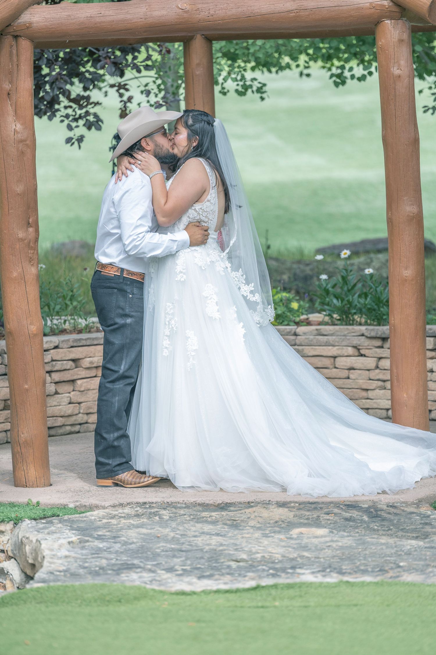 A romantic wedding couple shares a tender moment under a rustic wooden pergola in a garden setting.