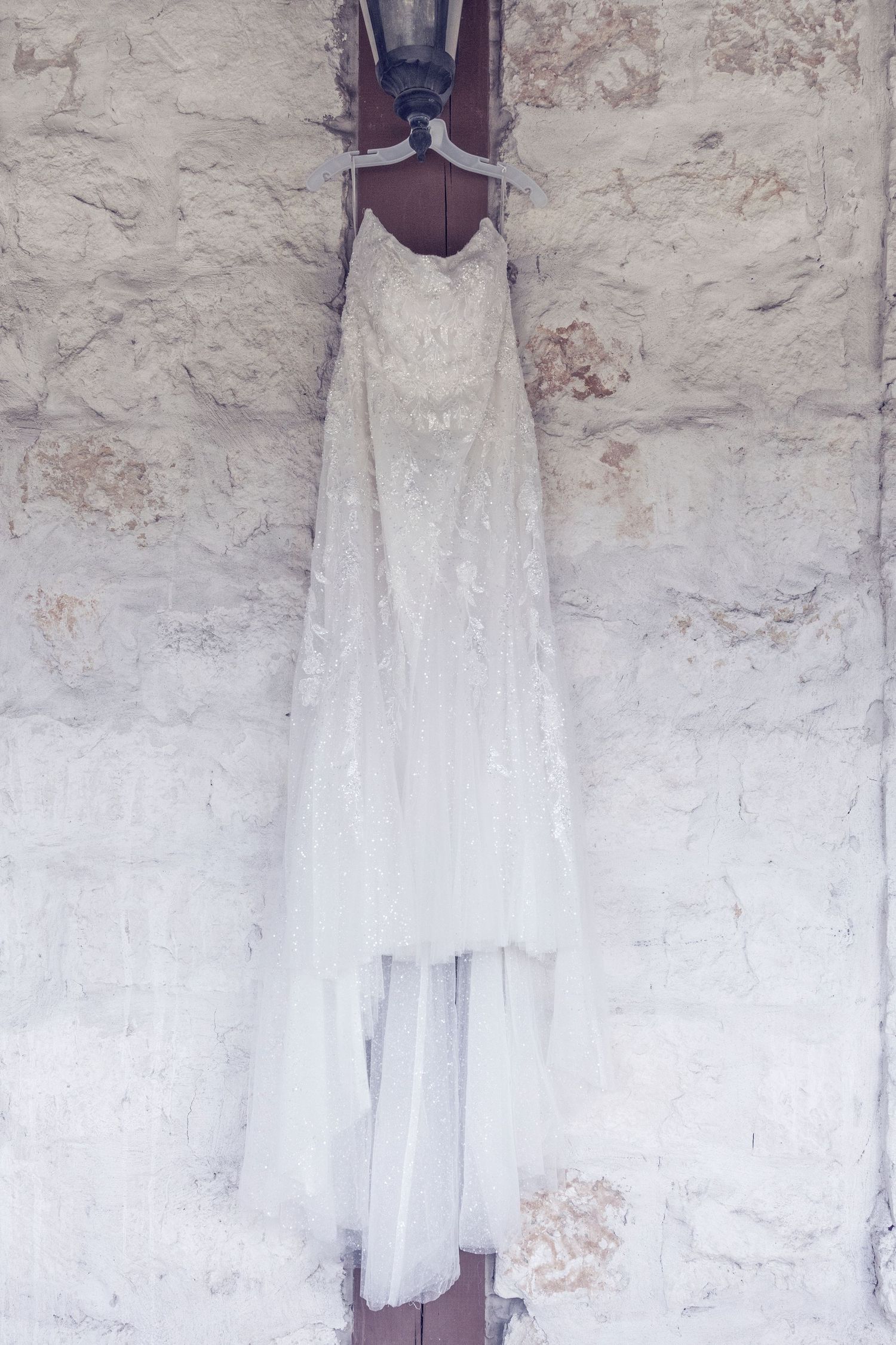 A vintage white lace dress hangs against a rustic stone wall, showcasing delicate details and romantic styling.