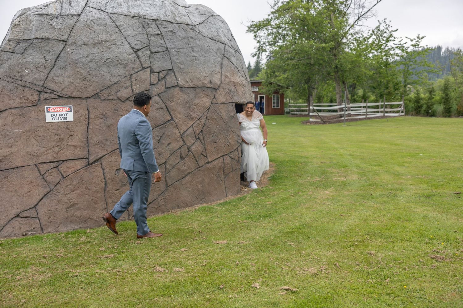 People explore the large outdoor climbing wall facility surrounded by green grass and trees on a cloudy day.