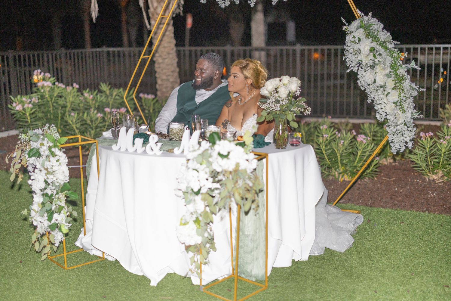A couple sits at an elegantly decorated sweetheart table with white linens and floral arrangements on an outdoor lawn.