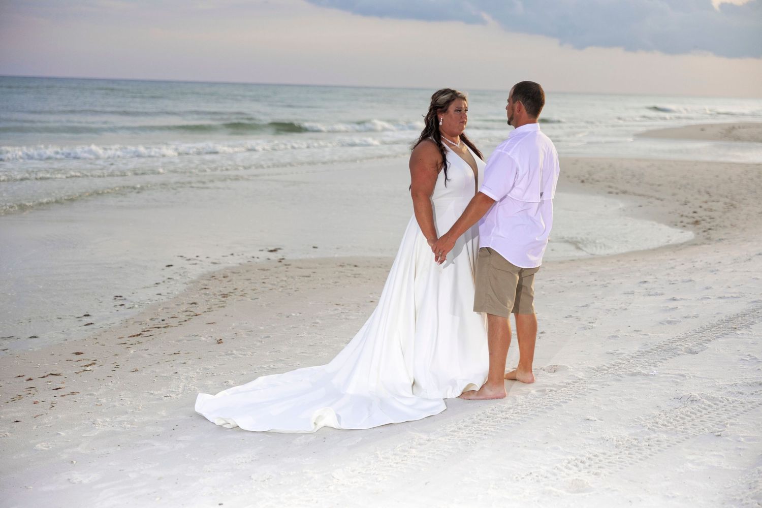 A couple in white attire shares romantic moments along a serene beach during sunset.