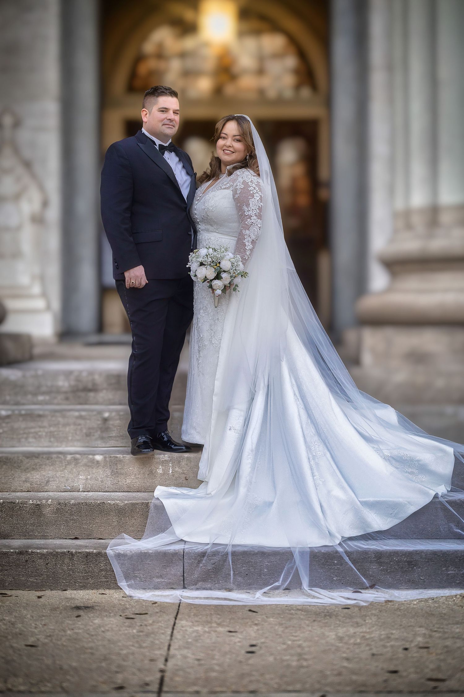 A romantic wedding couple poses on stone steps at a grand venue with the bride's white lace gown flowing beautifully.