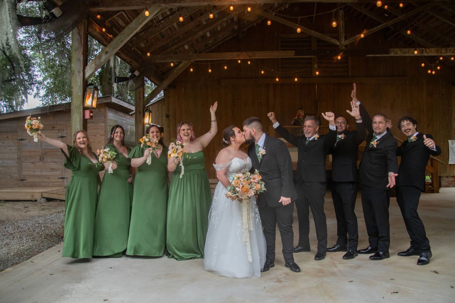 Wedding party poses together in a rustic barn venue decorated with twinkling string lights.