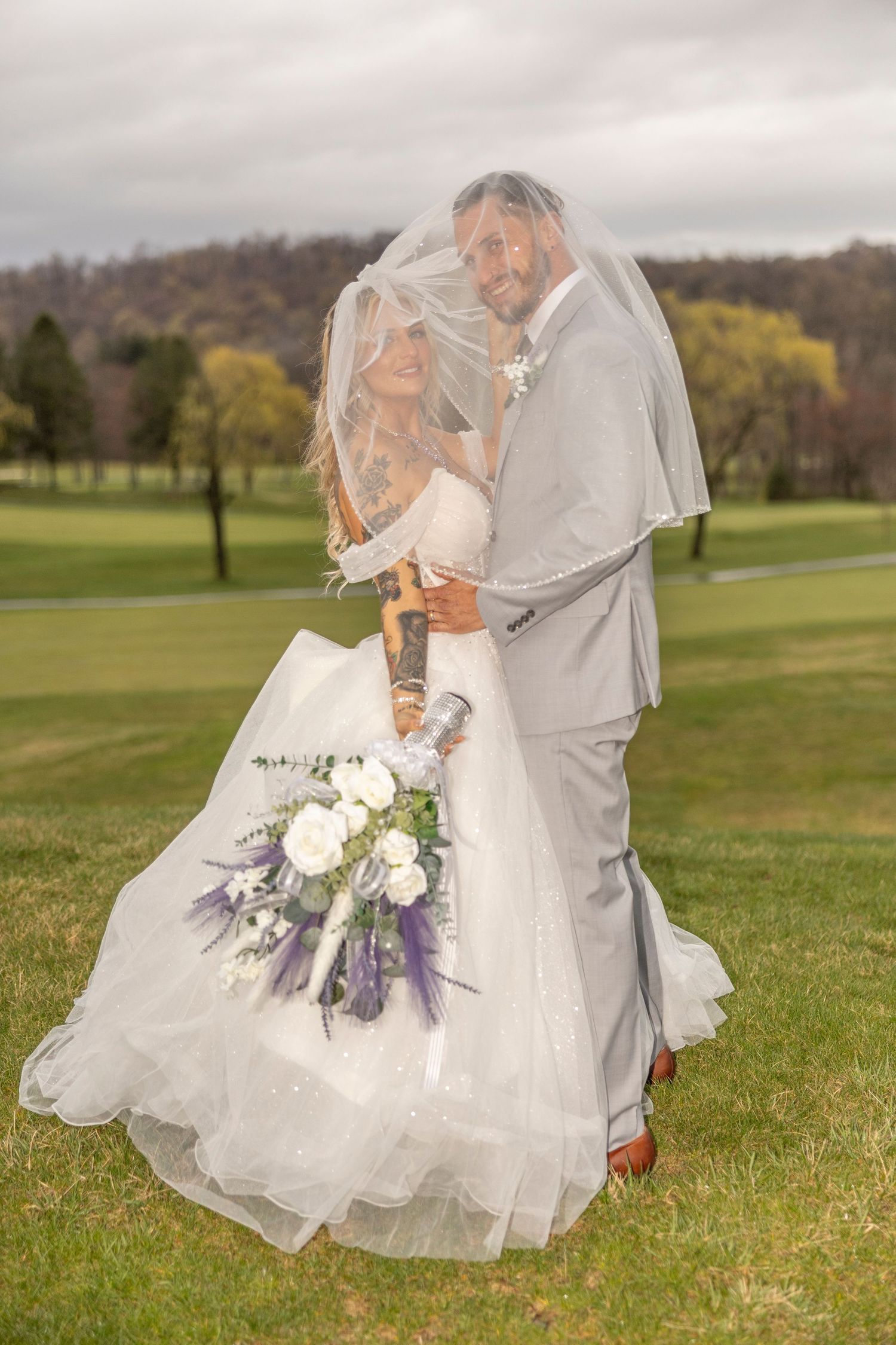 Romantic wedding photo of couple embracing under veil on golf course with autumn trees and purple bouquet.