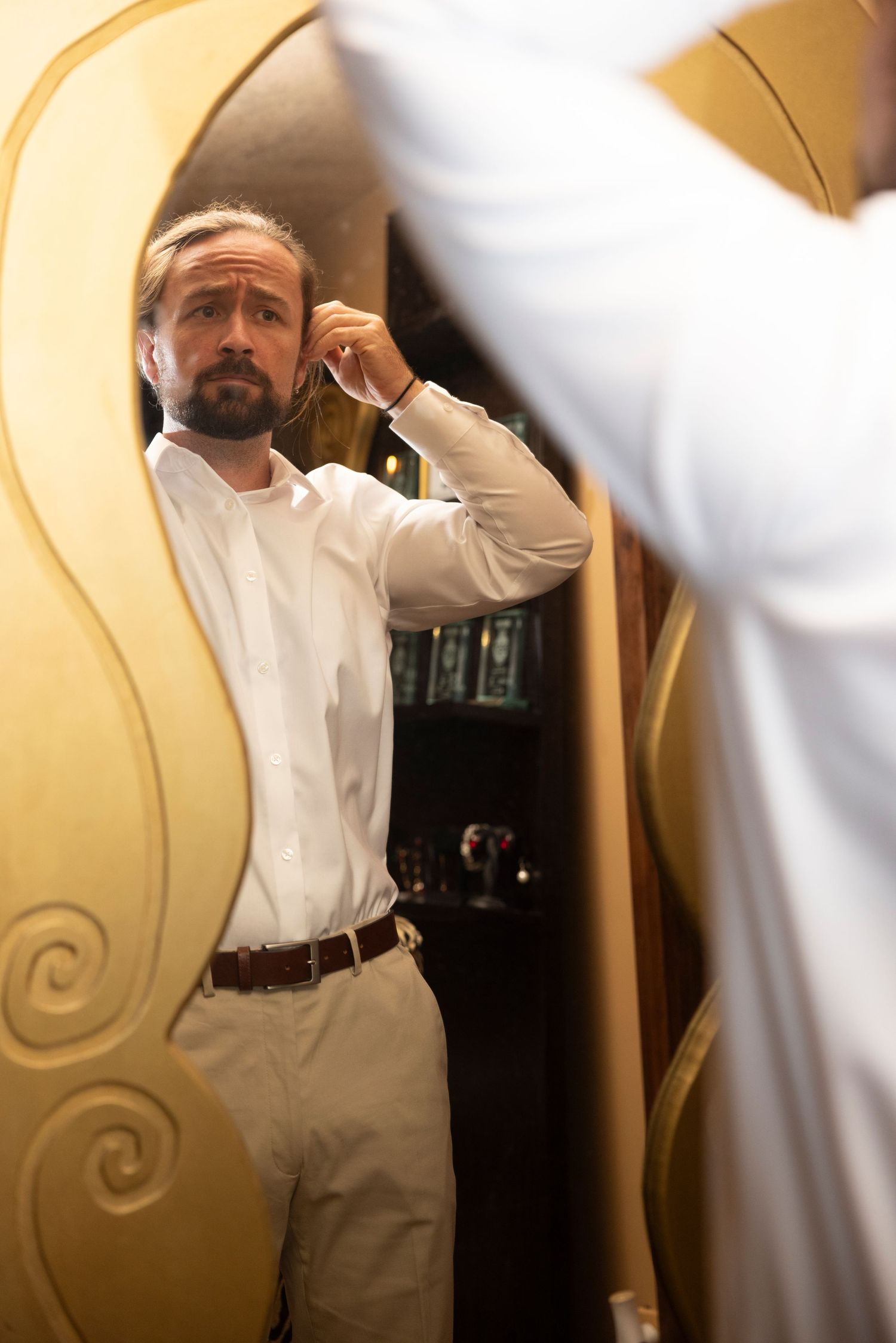 A man in white dress shirt adjusts his collar while looking in an ornate mirror.