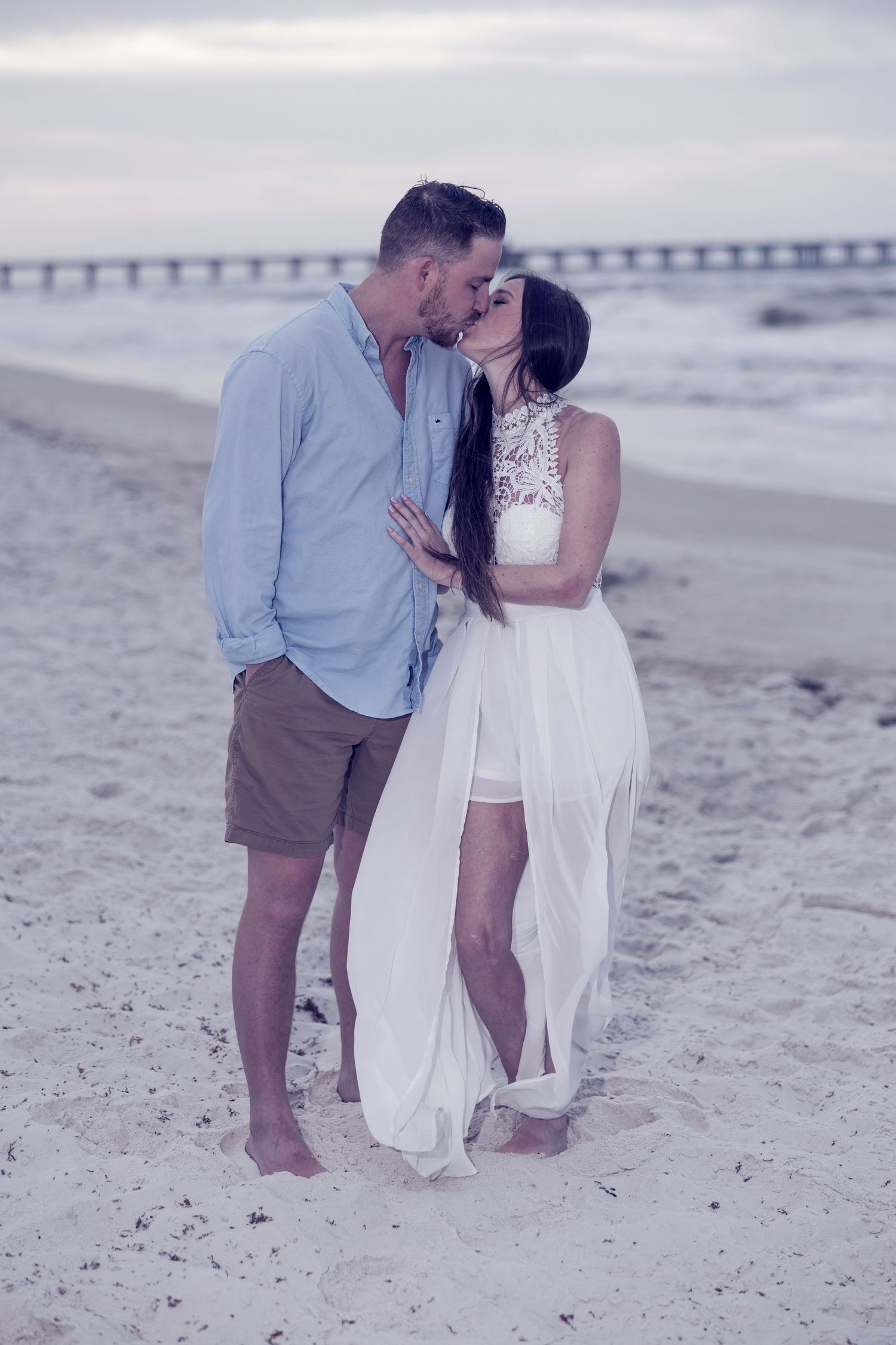 Couple sharing a romantic kiss on a sandy beach at dusk, woman in white flowing dress, man in blue shirt.