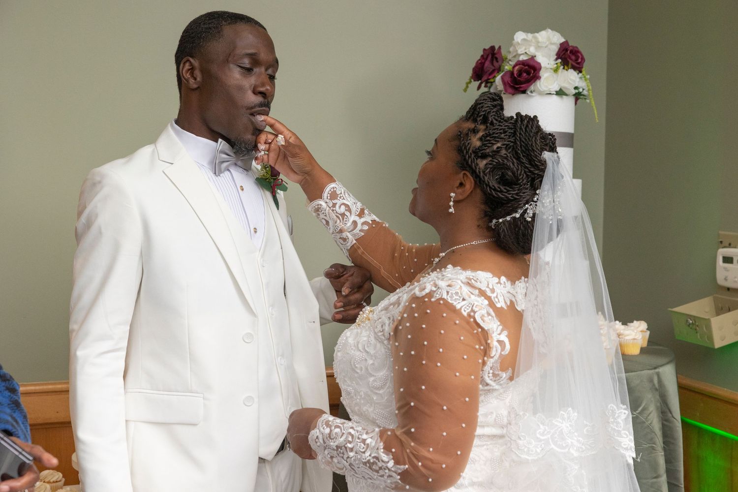 Bride in lace wedding gown feeds groom in white suit during wedding ceremony reception celebration.