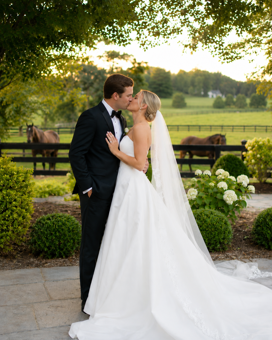 Bride and groom share a kiss in a white ballgown and tuxedo with horses and lush greenery in the background.