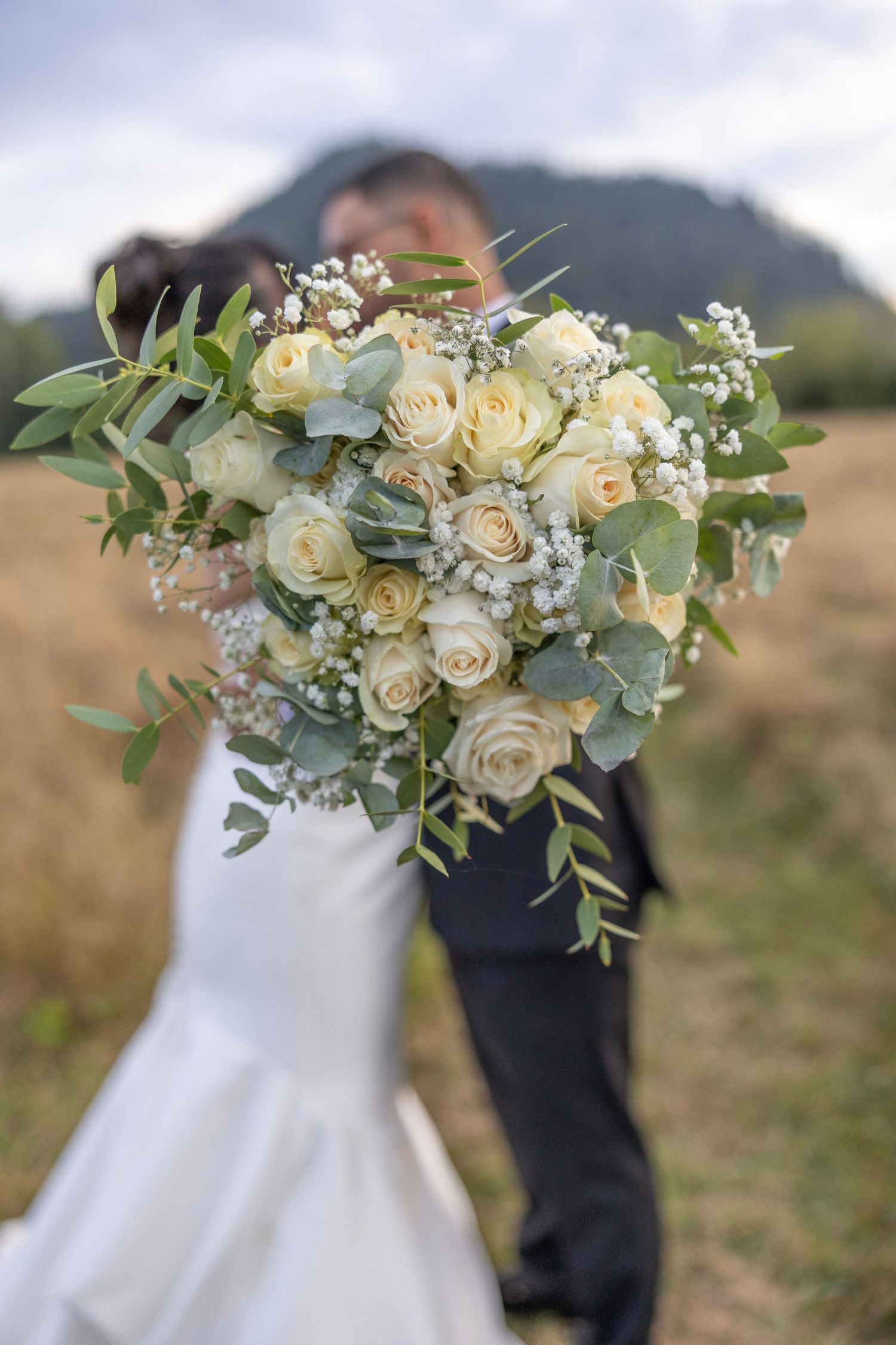 Elegant white and cream floral wedding bouquet with roses and greenery against a scenic mountain backdrop.