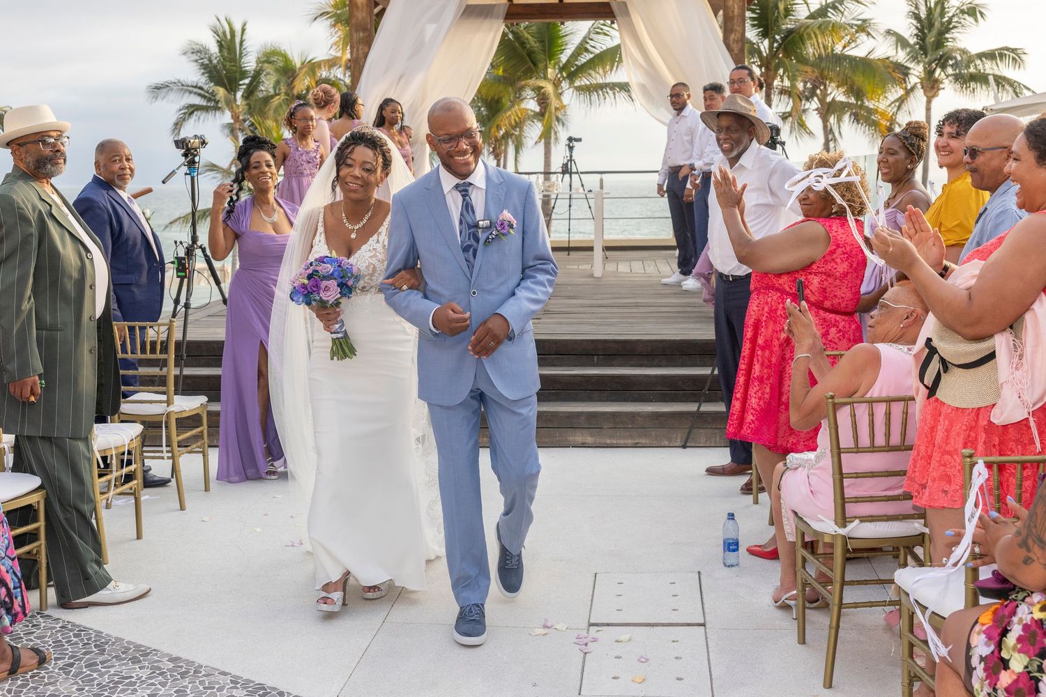 A joyful wedding ceremony at a tropical beach venue with guests in colorful attire lining the aisle.