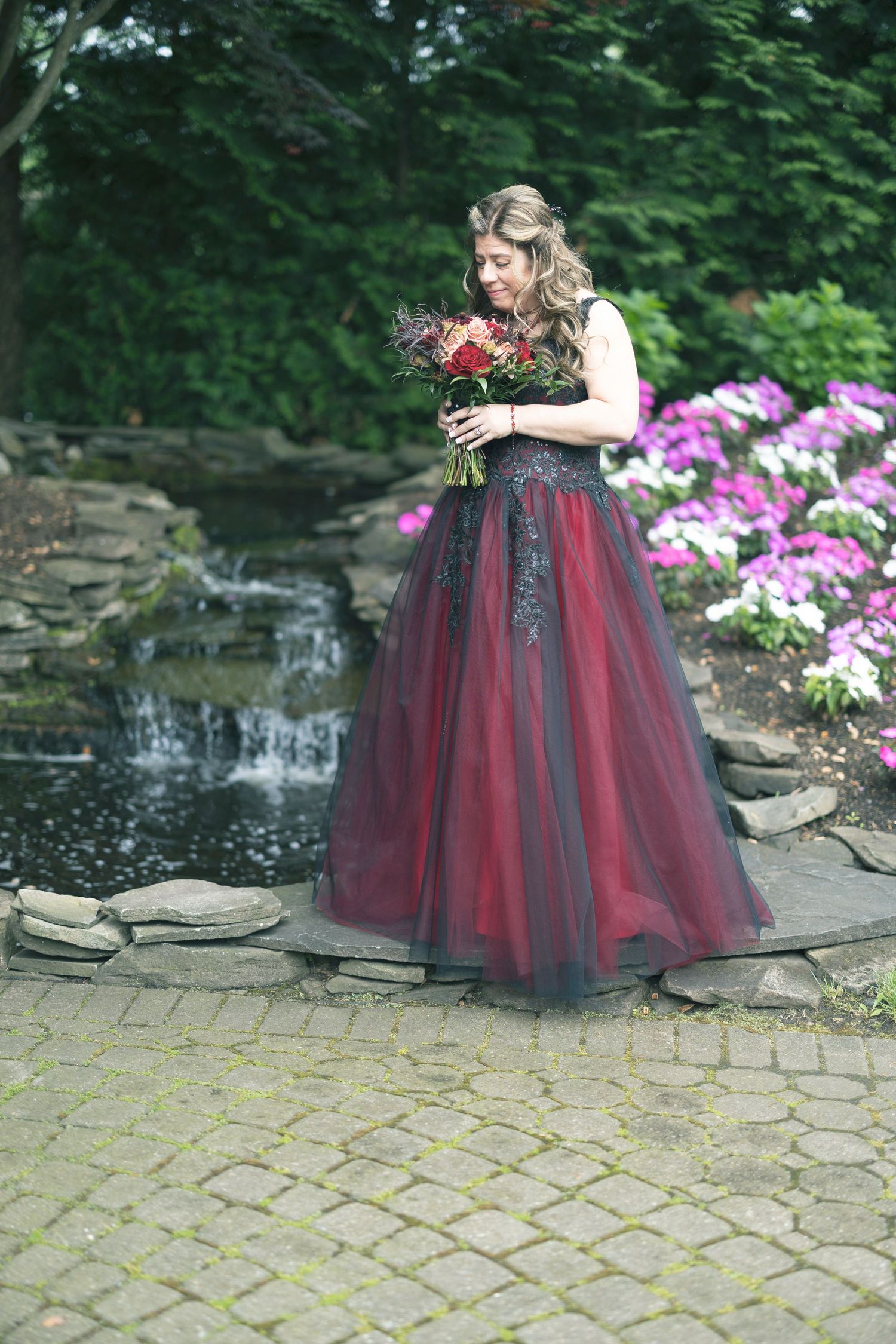A person in a burgundy and black tulle gown poses near a garden waterfall feature and pink flowers.
