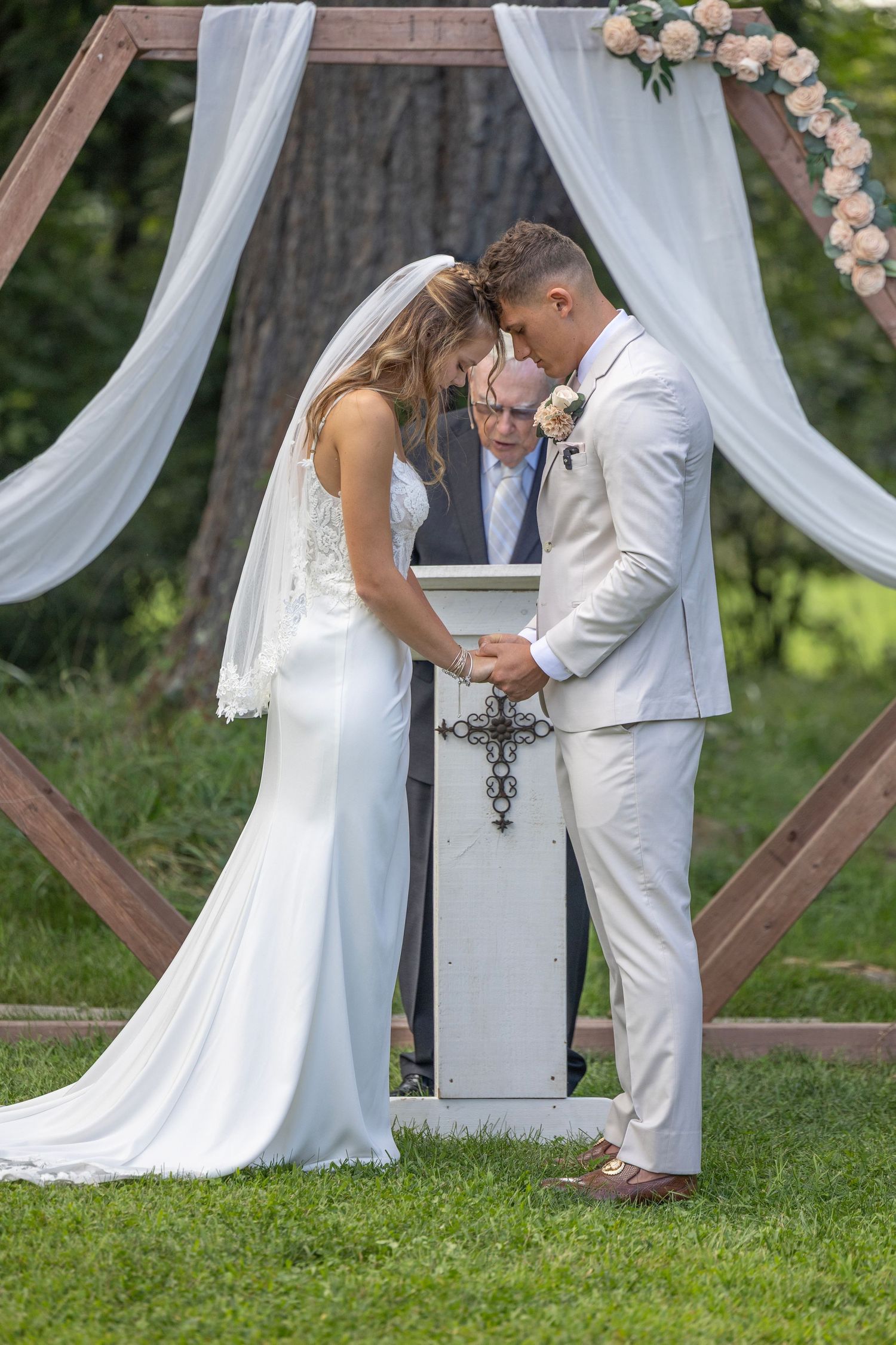 Couple performs unity cross ceremony during outdoor wedding with white draped arch and floral decorations.