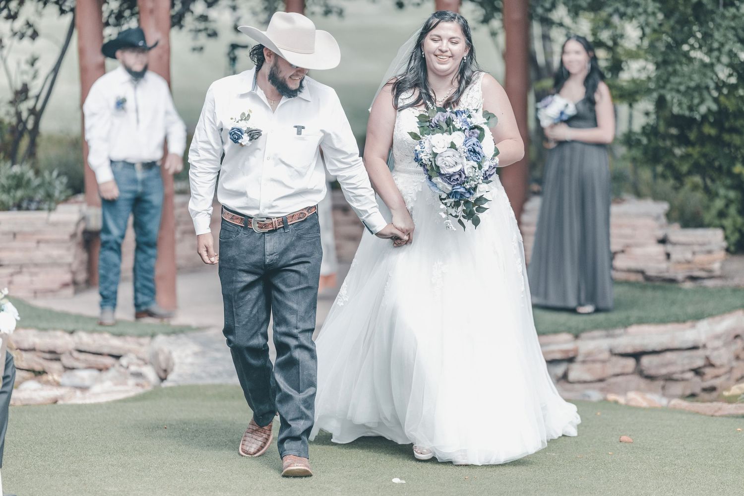 A couple walks down a grassy aisle during an outdoor wedding ceremony while guests look on in the background.
