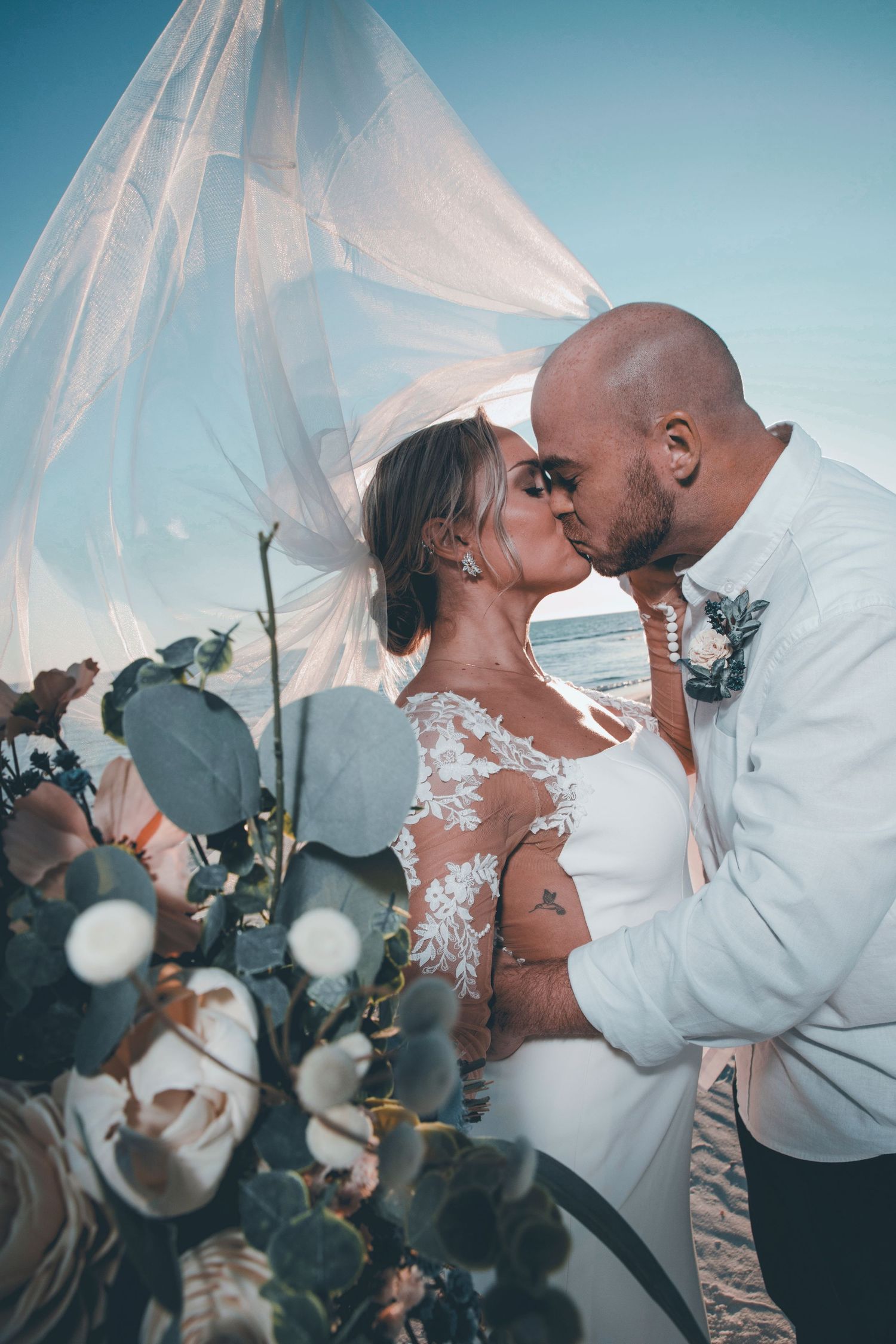A romantic couple shares an intimate kiss under a sheer white canopy with eucalyptus leaves in the background.