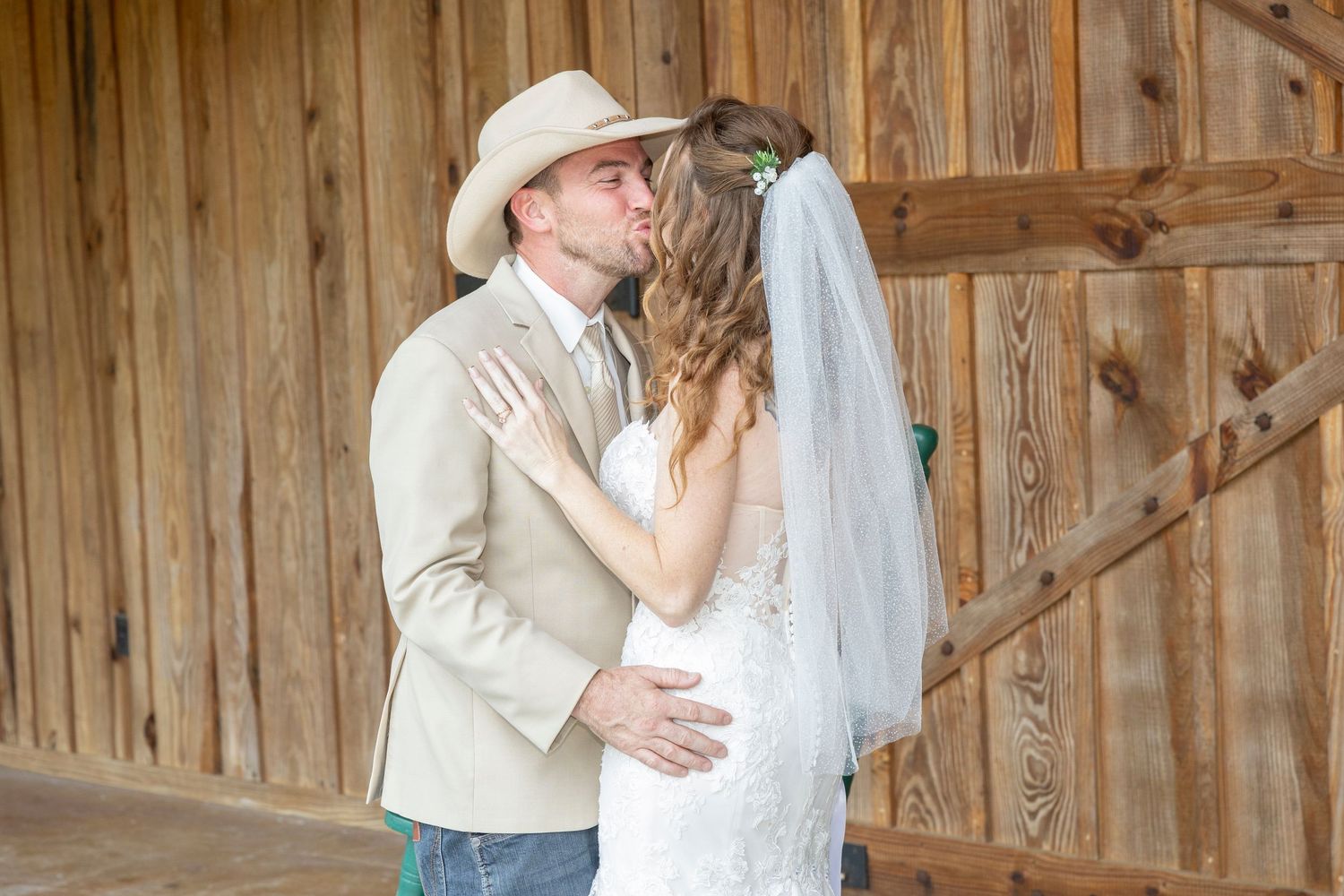 A couple in wedding attire embraces in front of a rustic wooden barn wall.