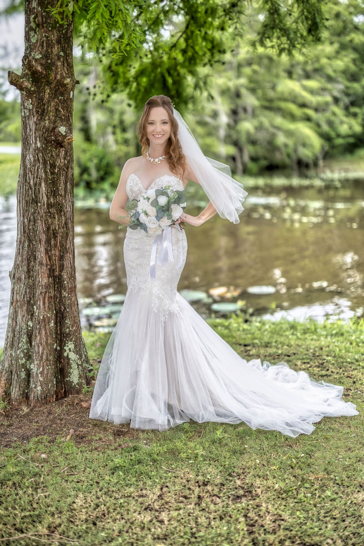 A bride in a white wedding dress poses gracefully near a pond under tall trees in a scenic outdoor setting.