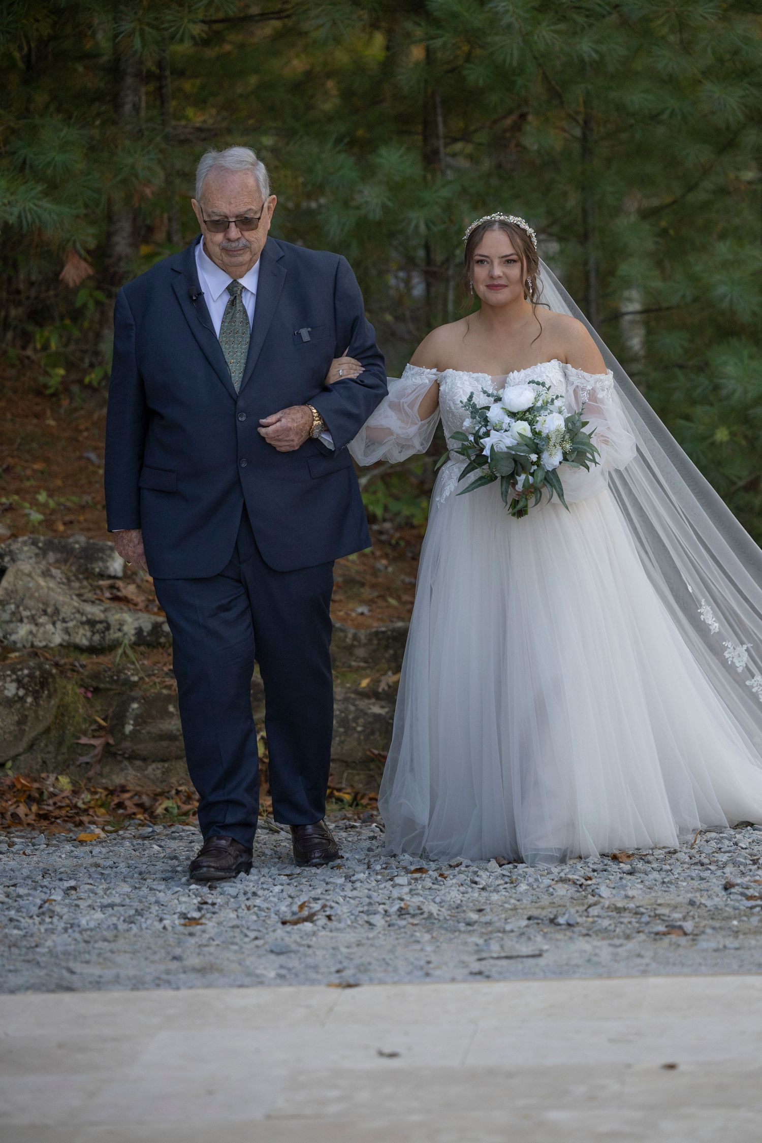 A bride in a white off-shoulder gown and long veil walks arm-in-arm with an escort on a gravel path in a wooded setting.