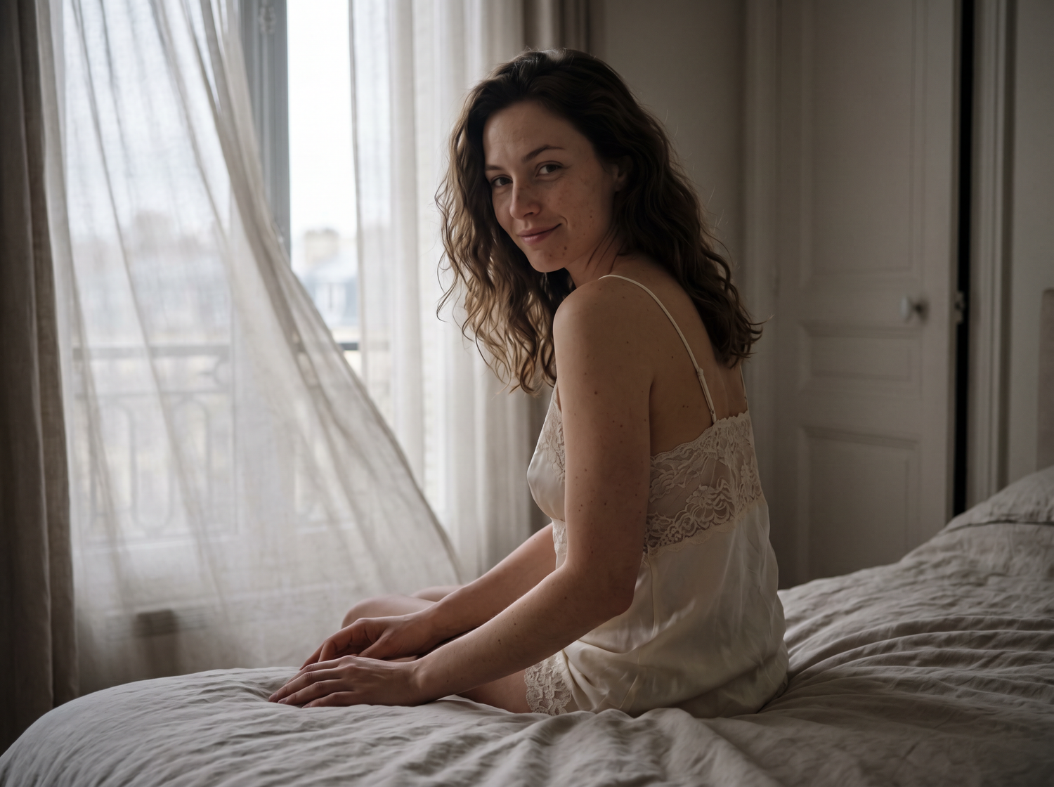 Young woman with curly hair sitting on bed in white slip dress by sheer curtained window.