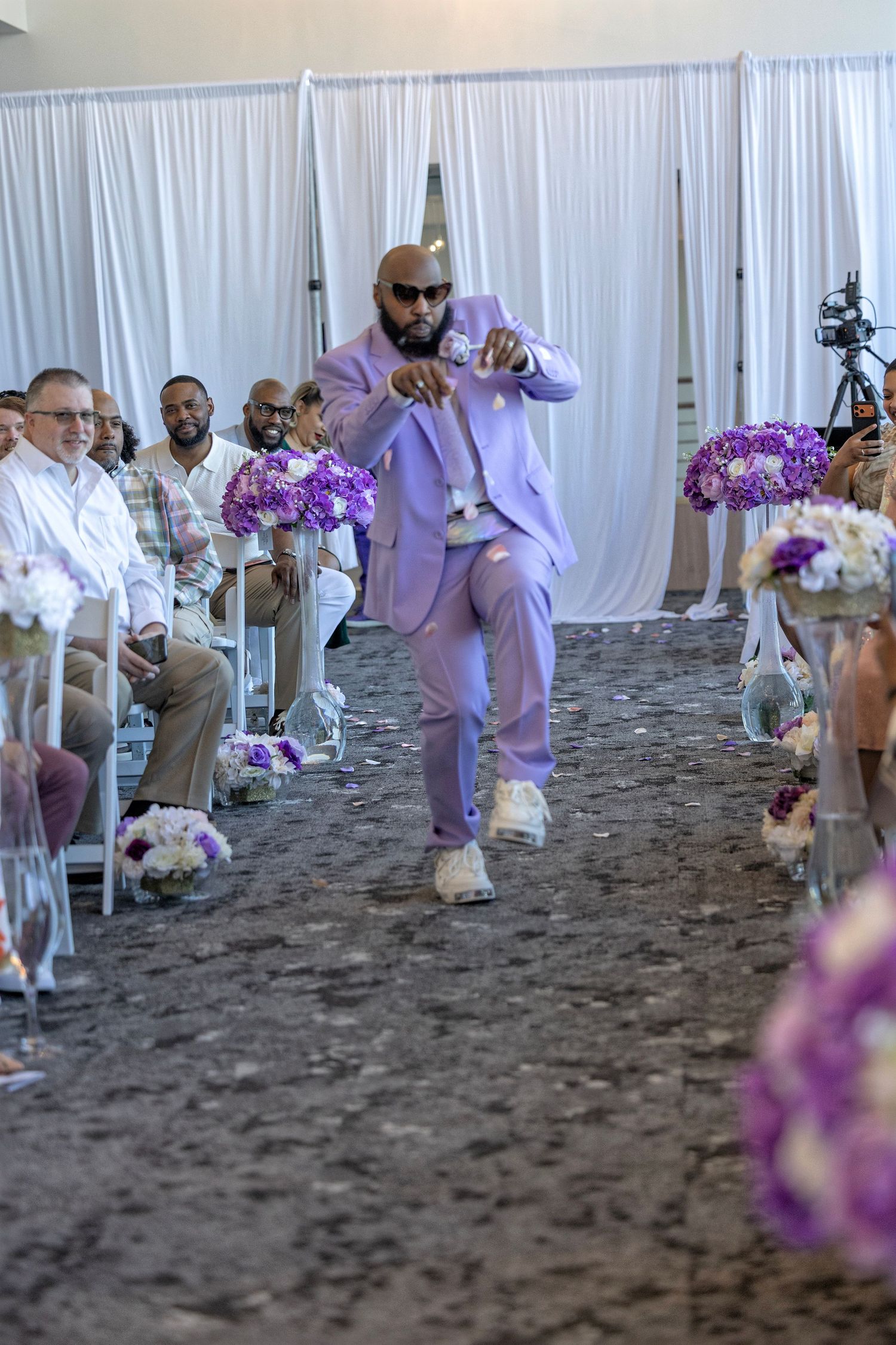 A man in a lavender suit dances down the aisle at a wedding ceremony decorated with purple floral arrangements.