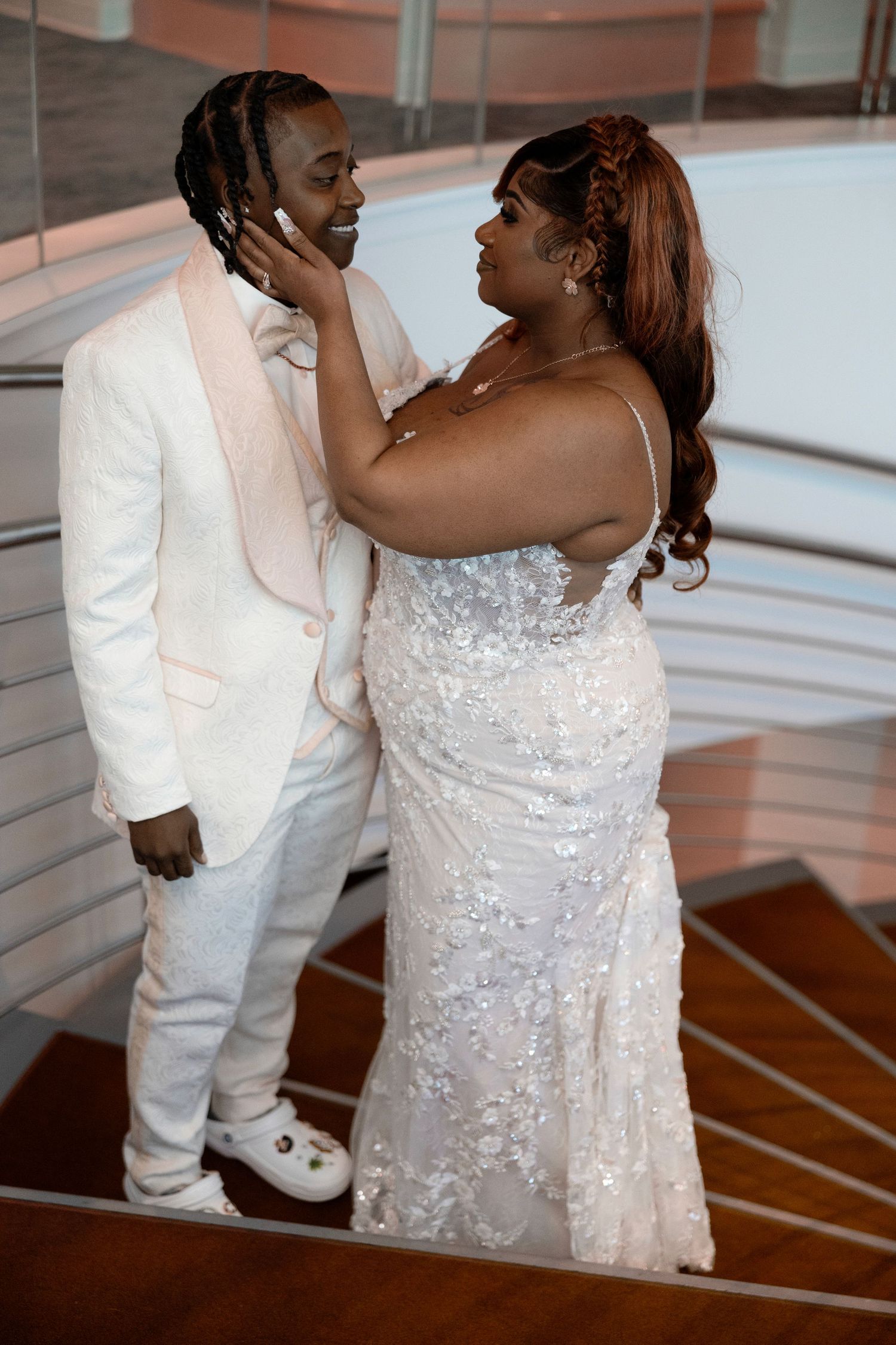 Couple in white wedding attire sharing a romantic moment on a staircase, bride in sequined gown touching groom's face.