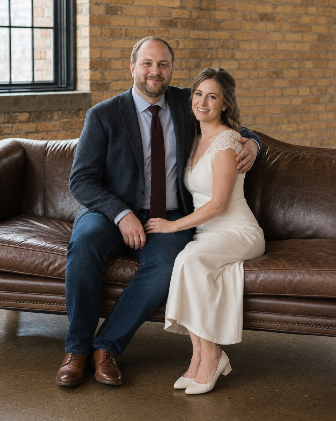 Couple in wedding attire sitting on leather couch, man in navy suit, woman in white dress, brick wall background.