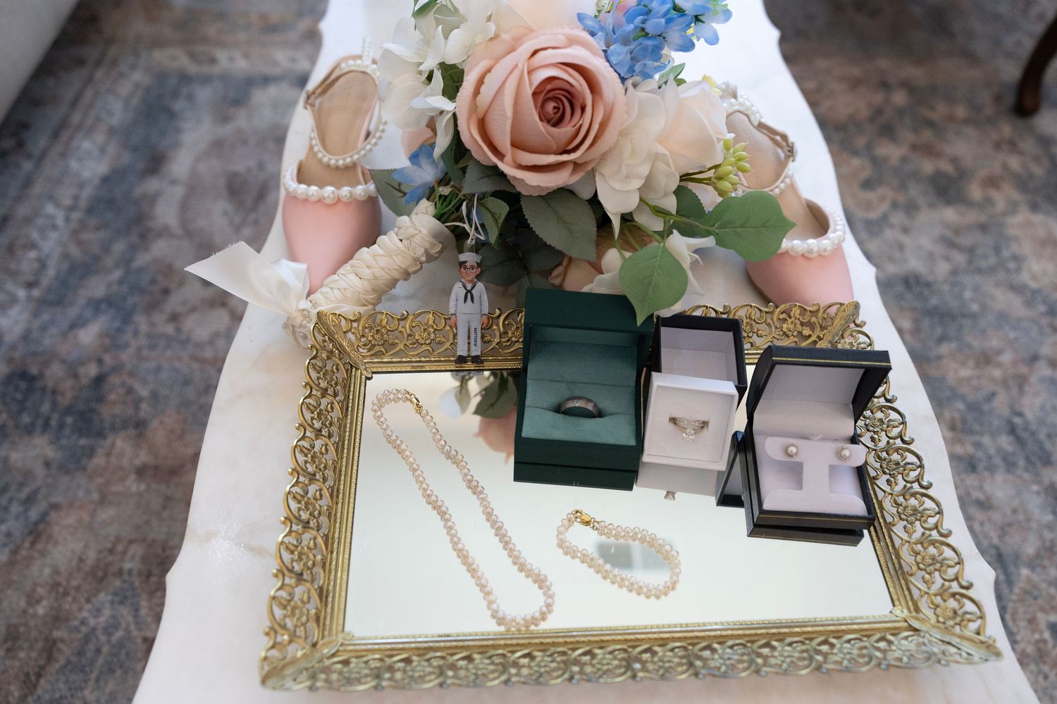 Gold tray displaying pearl necklace, engagement rings in boxes, and a pink rose bridal bouquet on a white table.