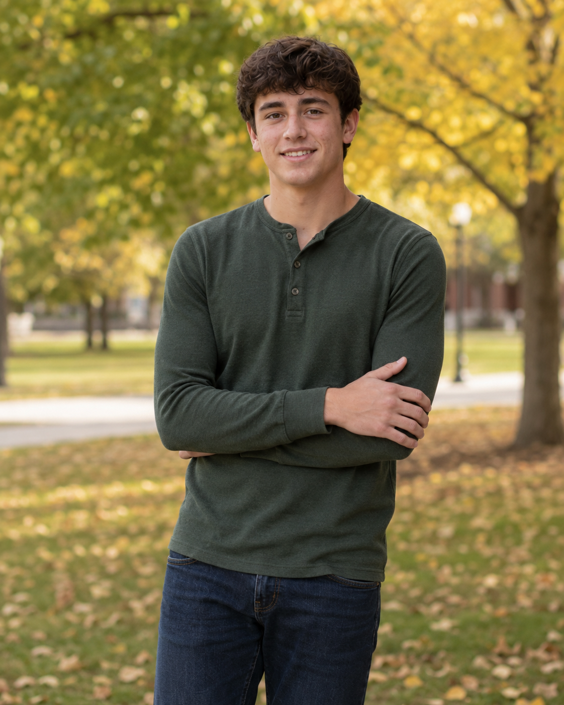Teen boy in olive green henley stands with arms crossed outdoors among fall foliage for senior photos.