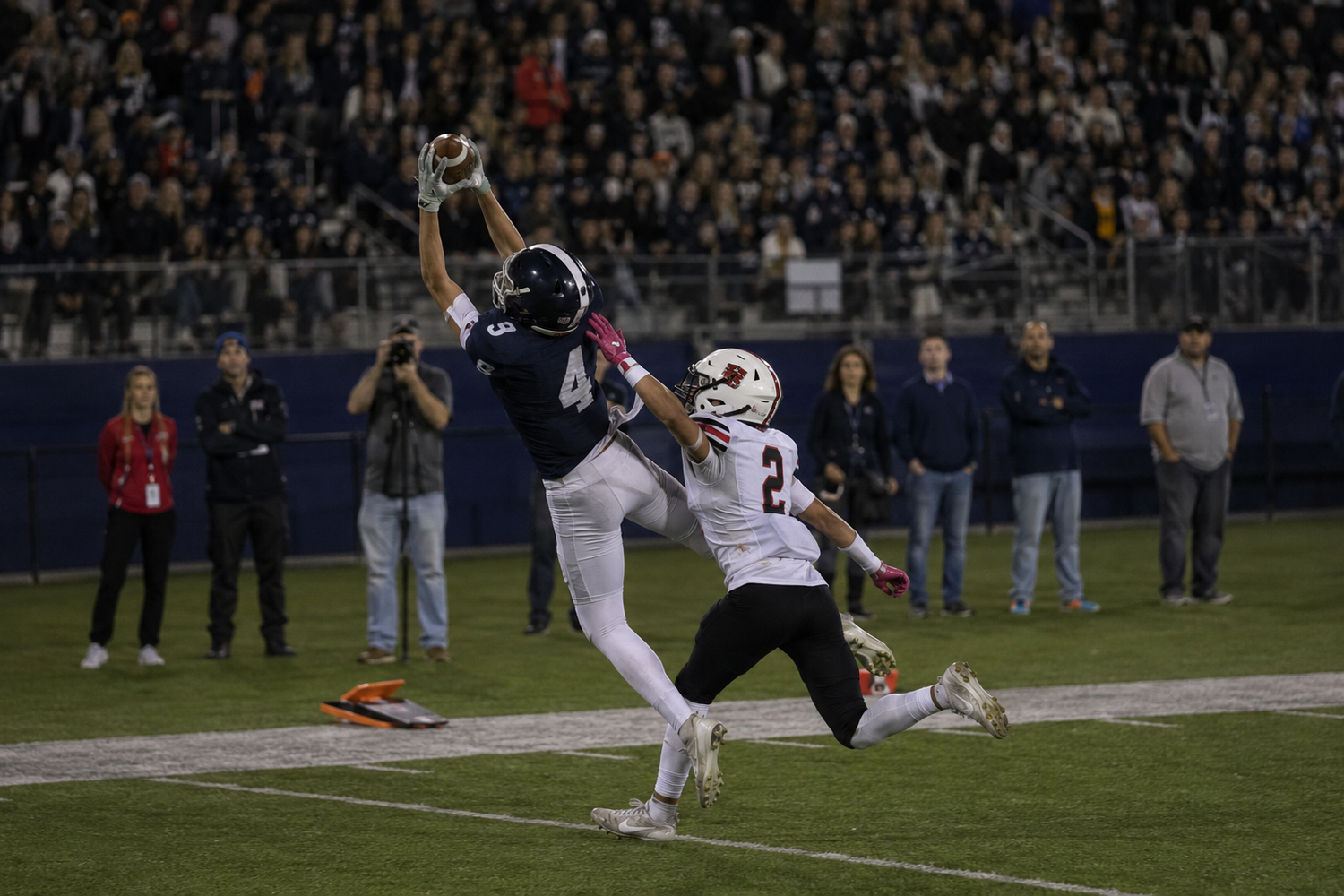 Football player catches pass in end zone while defender attempts coverage during night game.