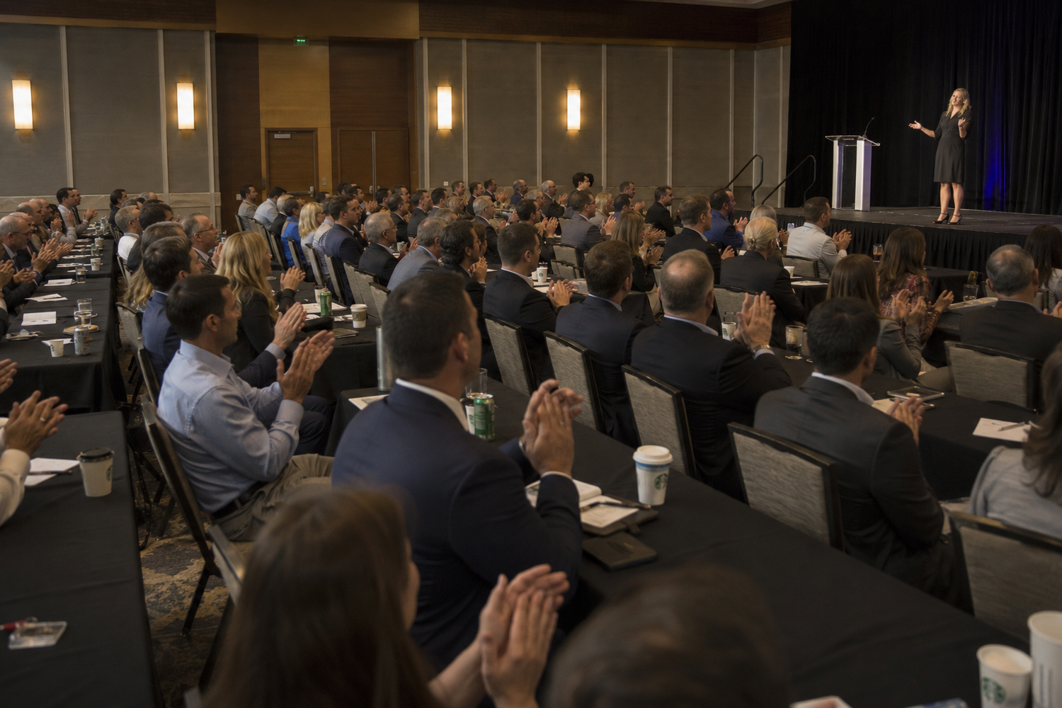 Large audience applauding a female speaker at a corporate conference in a modern hotel ballroom.
