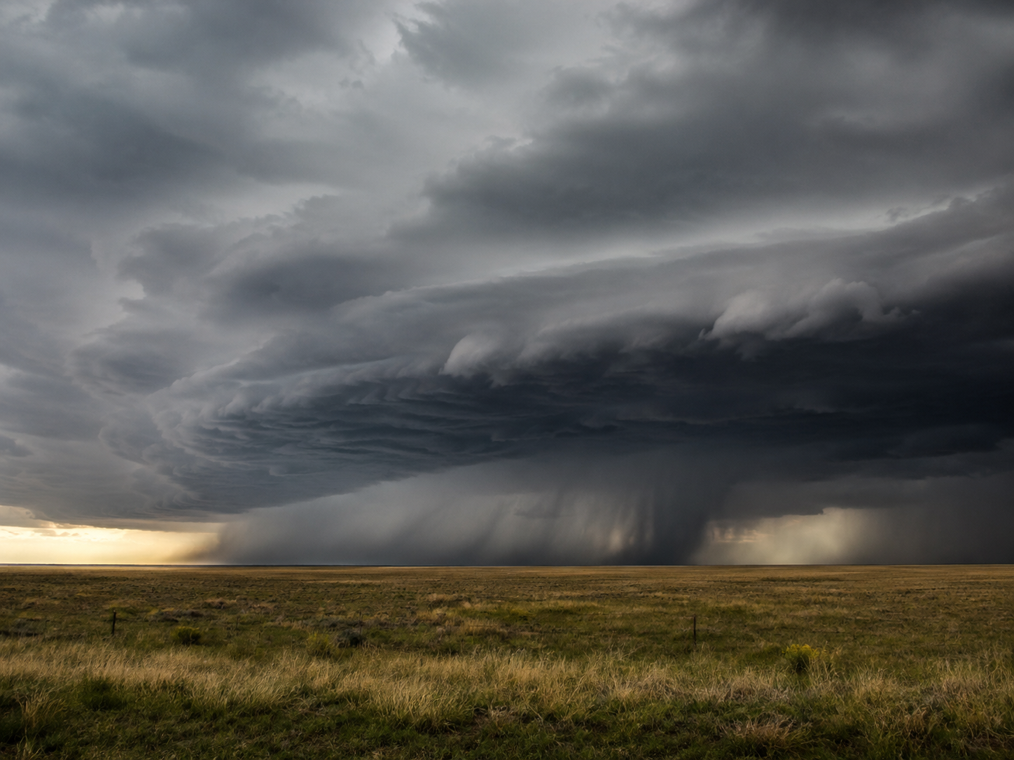 Massive supercell thunderstorm over flat grassland plains with dramatic swirling dark clouds and heavy rain curtain.