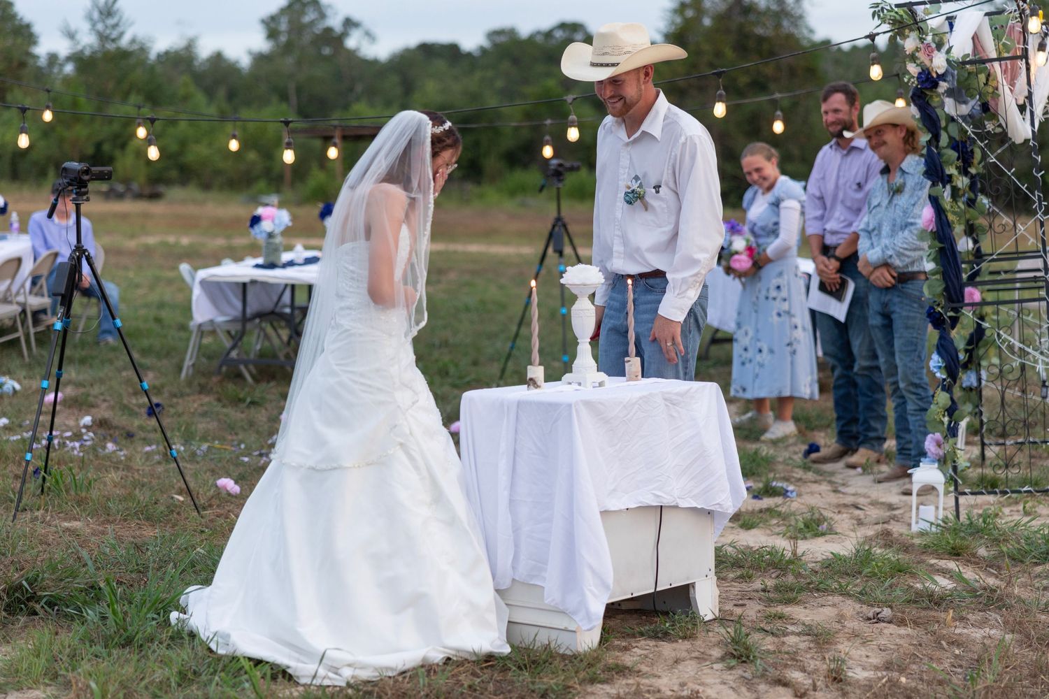 Outdoor rustic wedding ceremony with bride in white gown and veil, groom in cowboy hat, string lights and floral arch.