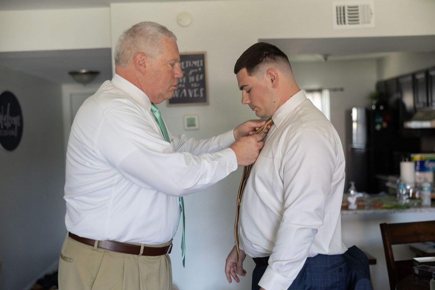 Older man helping groom adjust tie before wedding ceremony indoors.