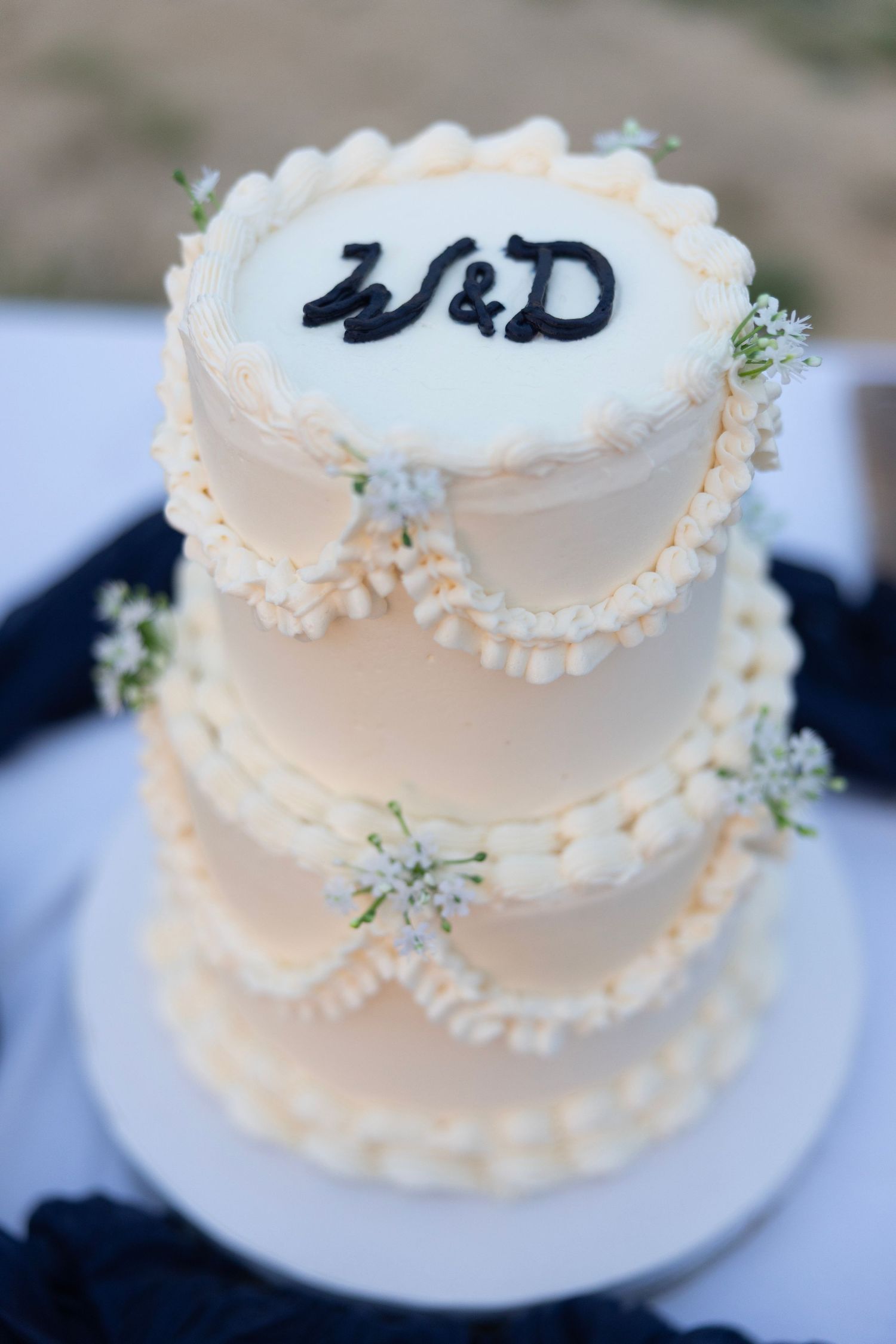 Two-tier white wedding cake with W&D monogram, pearl borders, and delicate white flowers on a white cake stand.