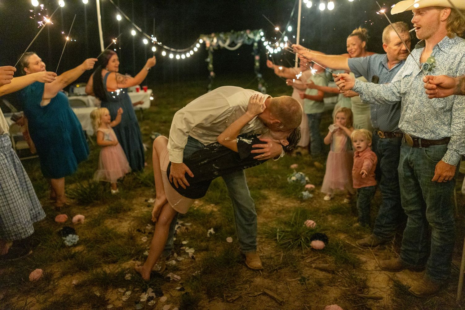 Couple sharing romantic dip kiss at outdoor wedding reception with sparklers and guests celebrating at night.
