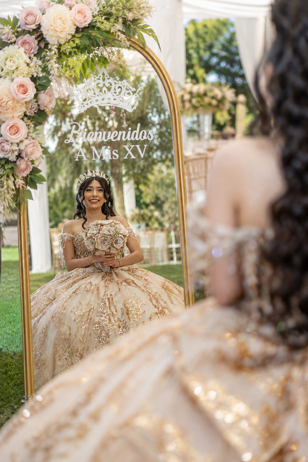 Young woman in gold quinceañera gown poses before floral arch mirror reading Bienvenidos A Mis XV.