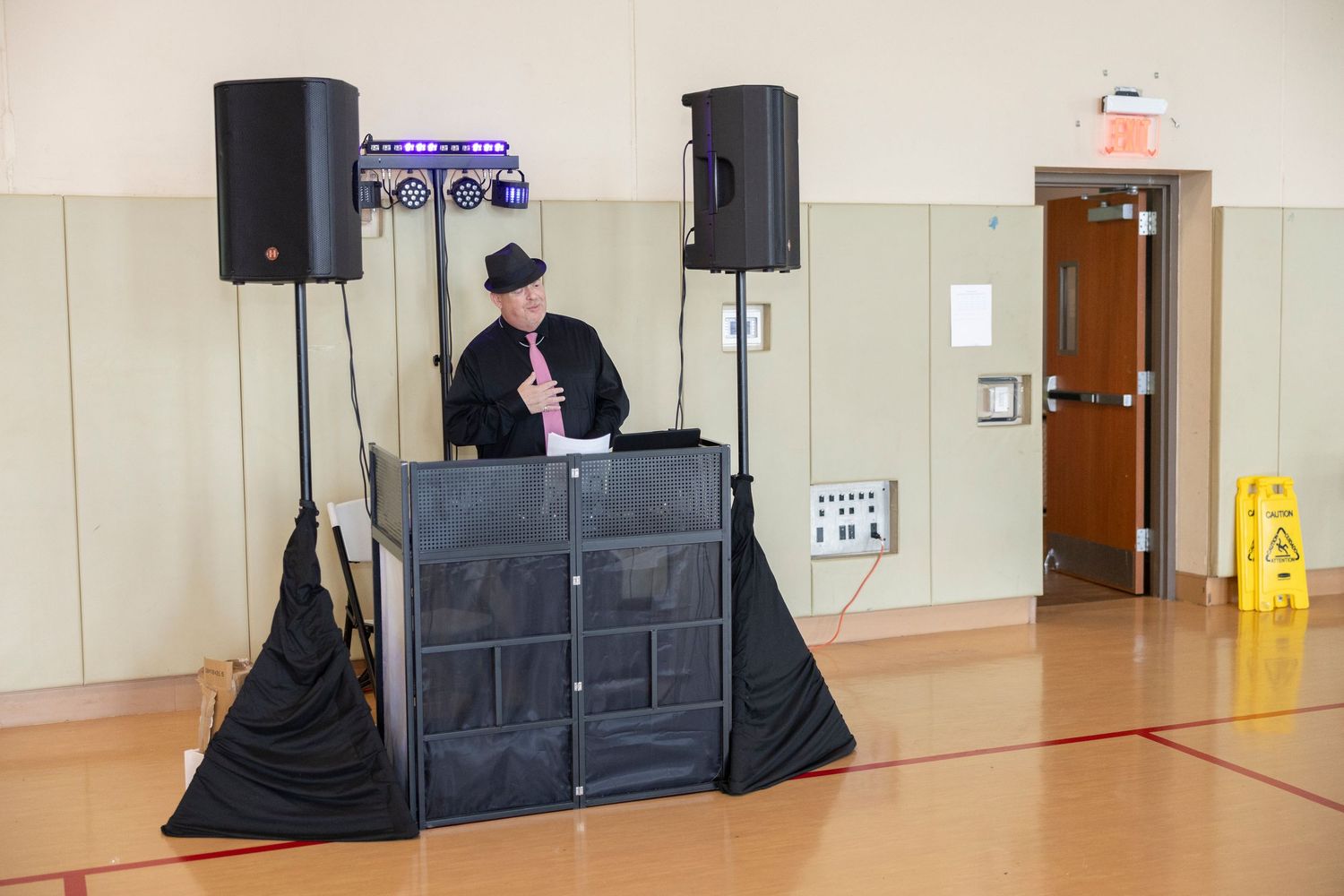 A DJ in a black hat performs behind a booth with speakers in a school gymnasium near a wet floor sign.
