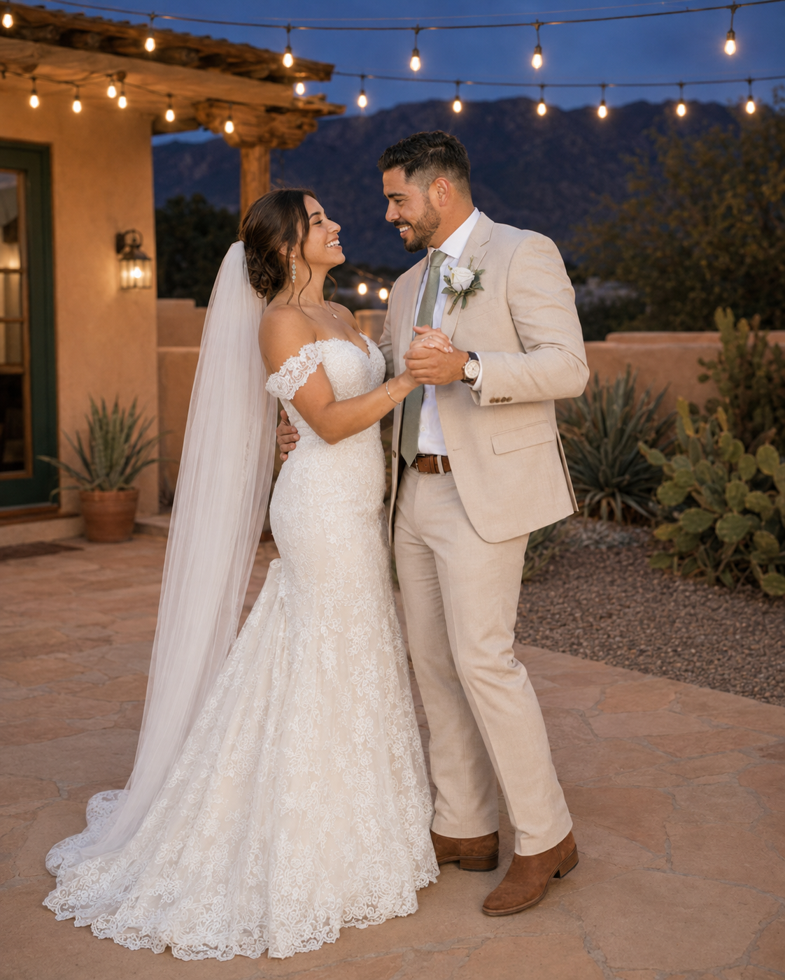 Bride in lace mermaid gown with veil and groom in tan suit share romantic moment at desert wedding venue at dusk.