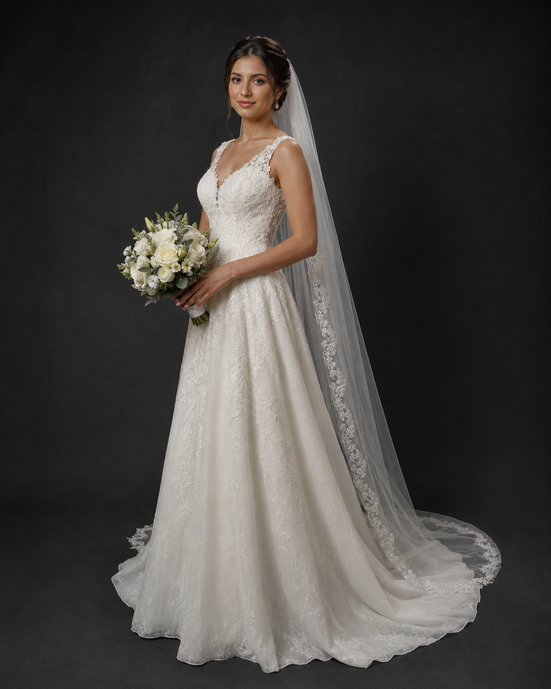 Bride in ivory lace A-line wedding gown with V-neck, long veil, holding white floral bouquet against dark background.