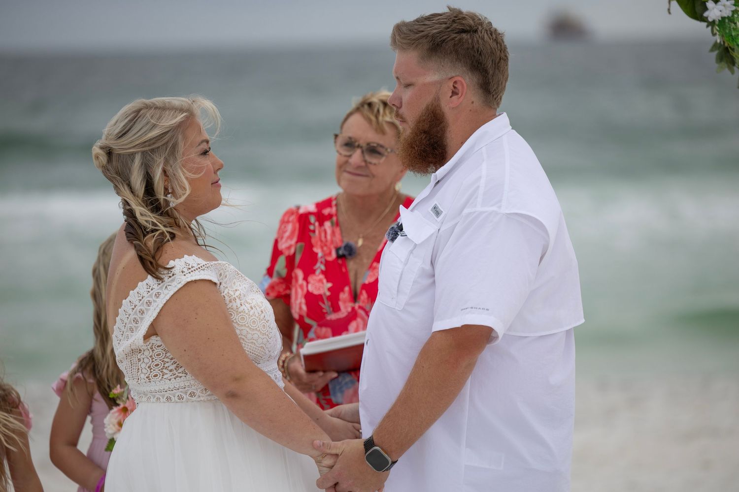 Couple exchanges vows on a sunny beach wedding ceremony with officiant in red dress, ocean waves in background.