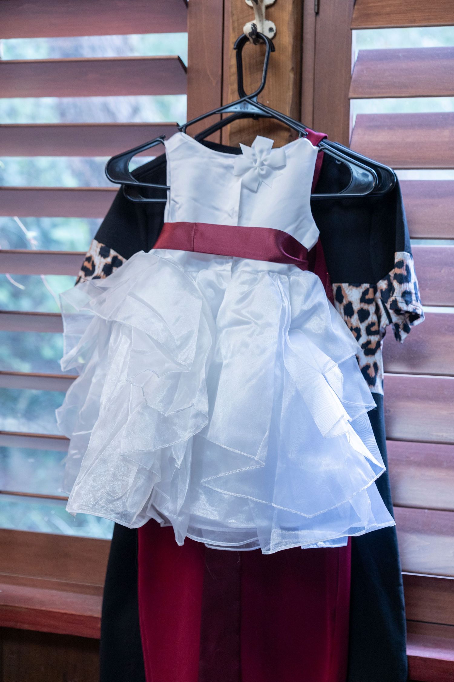 A white ruffled flower girl dress with burgundy sash hangs on display.