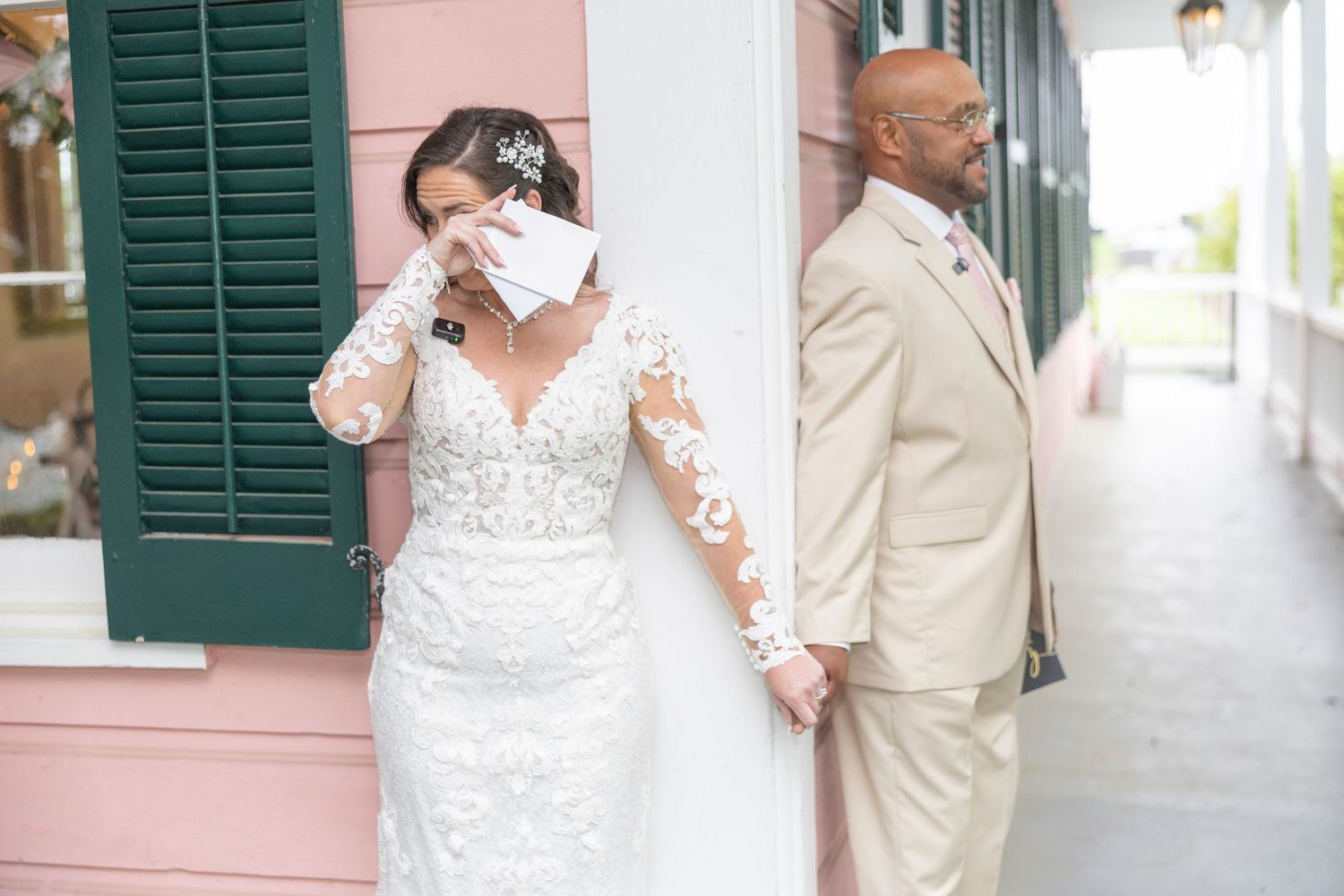 A wedding couple stands against a pink wall with green shutters in a first look moment before their ceremony.