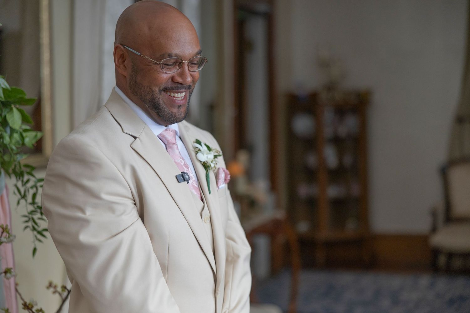 A joyful person wearing a cream suit with pink tie and boutonniere smiles warmly during a special occasion.