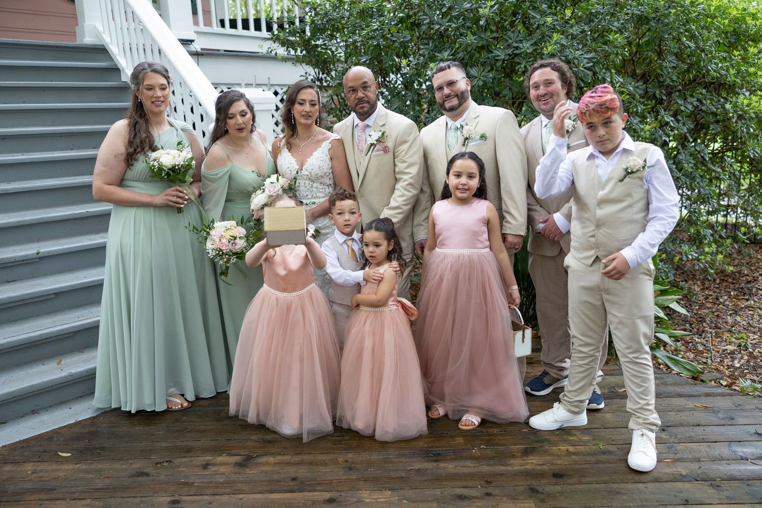 Wedding party members in pastel pink and sage green dresses pose with groomsmen in light tan suits outdoors.