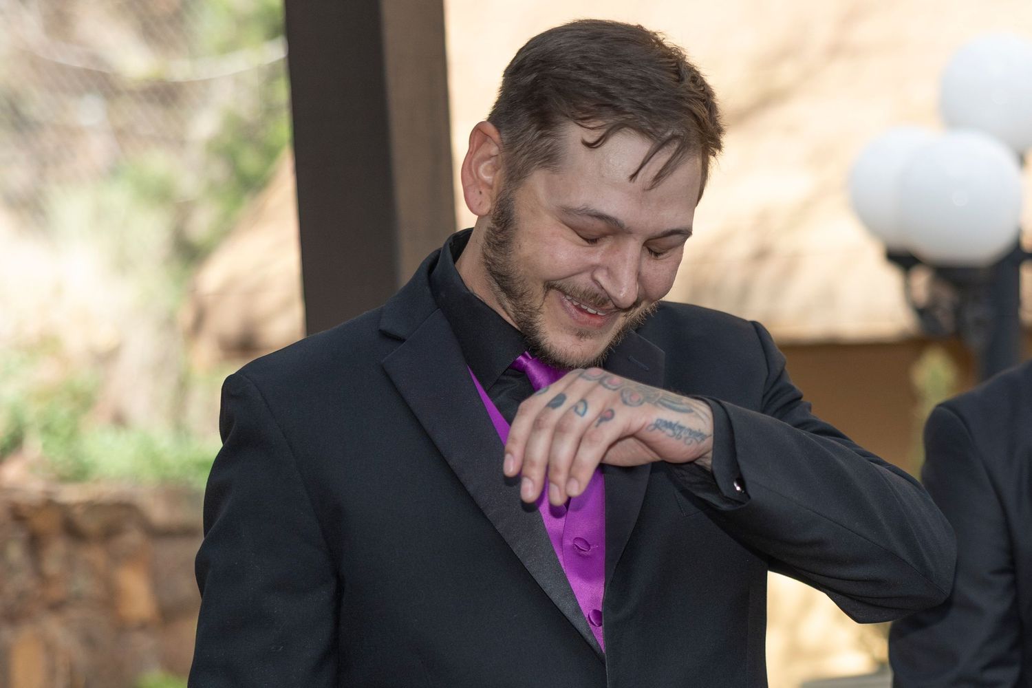 A person in a black suit and purple shirt laughs while checking their watch at an outdoor event.