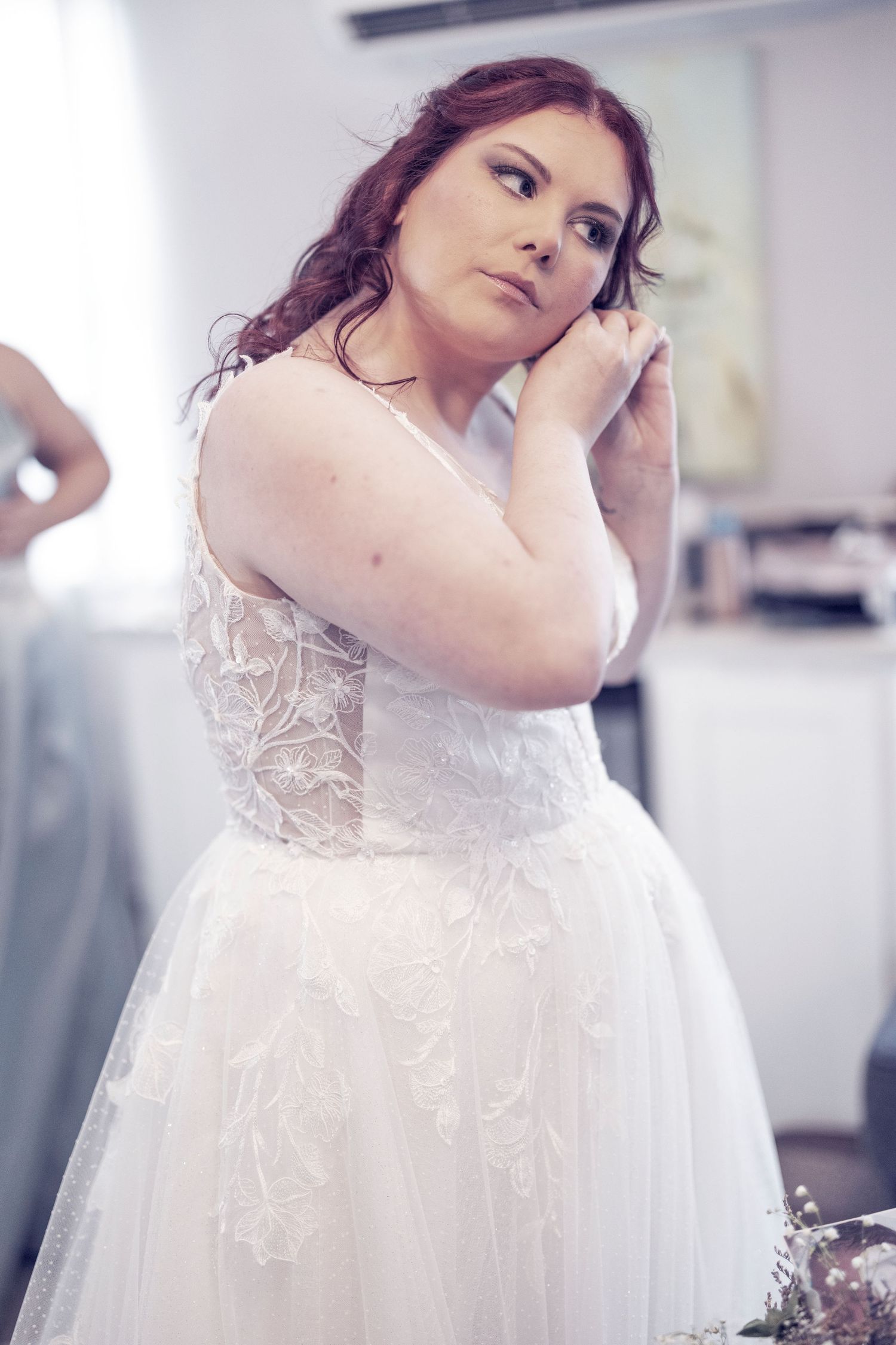 A bride in a white lace and tulle wedding gown poses during a fitting session in a bridal boutique.
