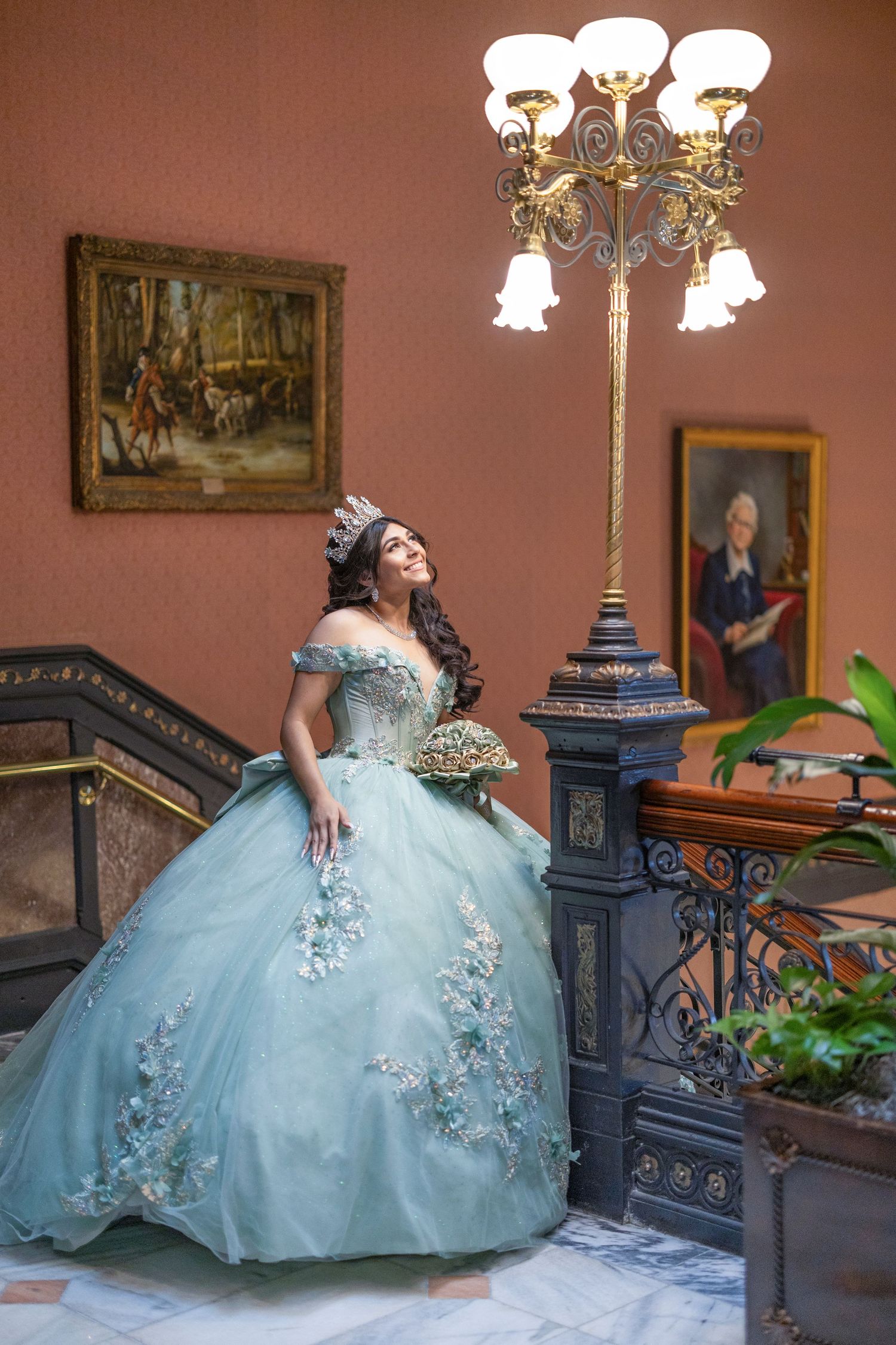 A person in a light blue quinceañera gown with floral embellishments poses on ornate stairs in an elegant indoor setting.