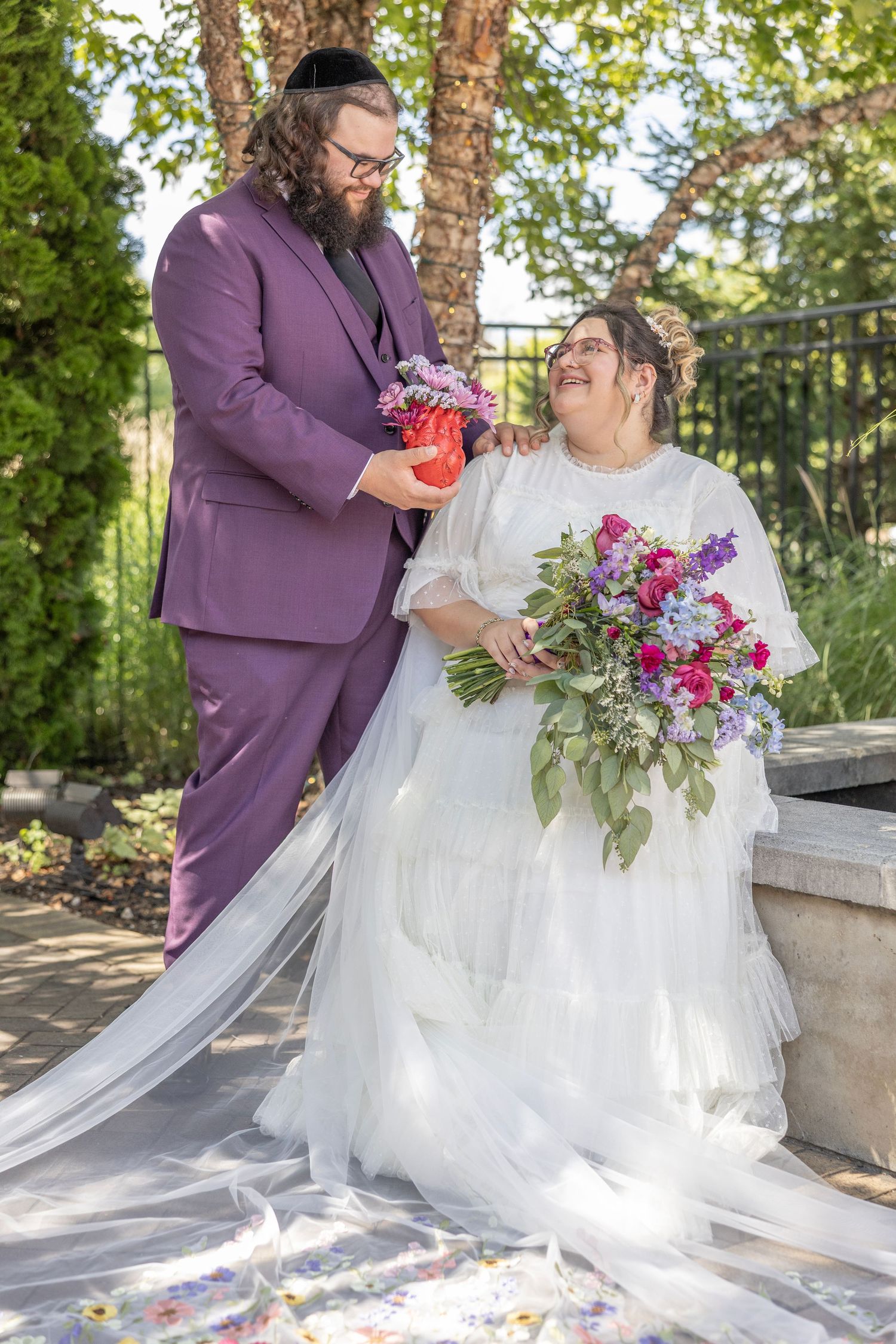 A couple shares a joyful moment in their wedding attire, with the bride in white and groom in purple outdoors.