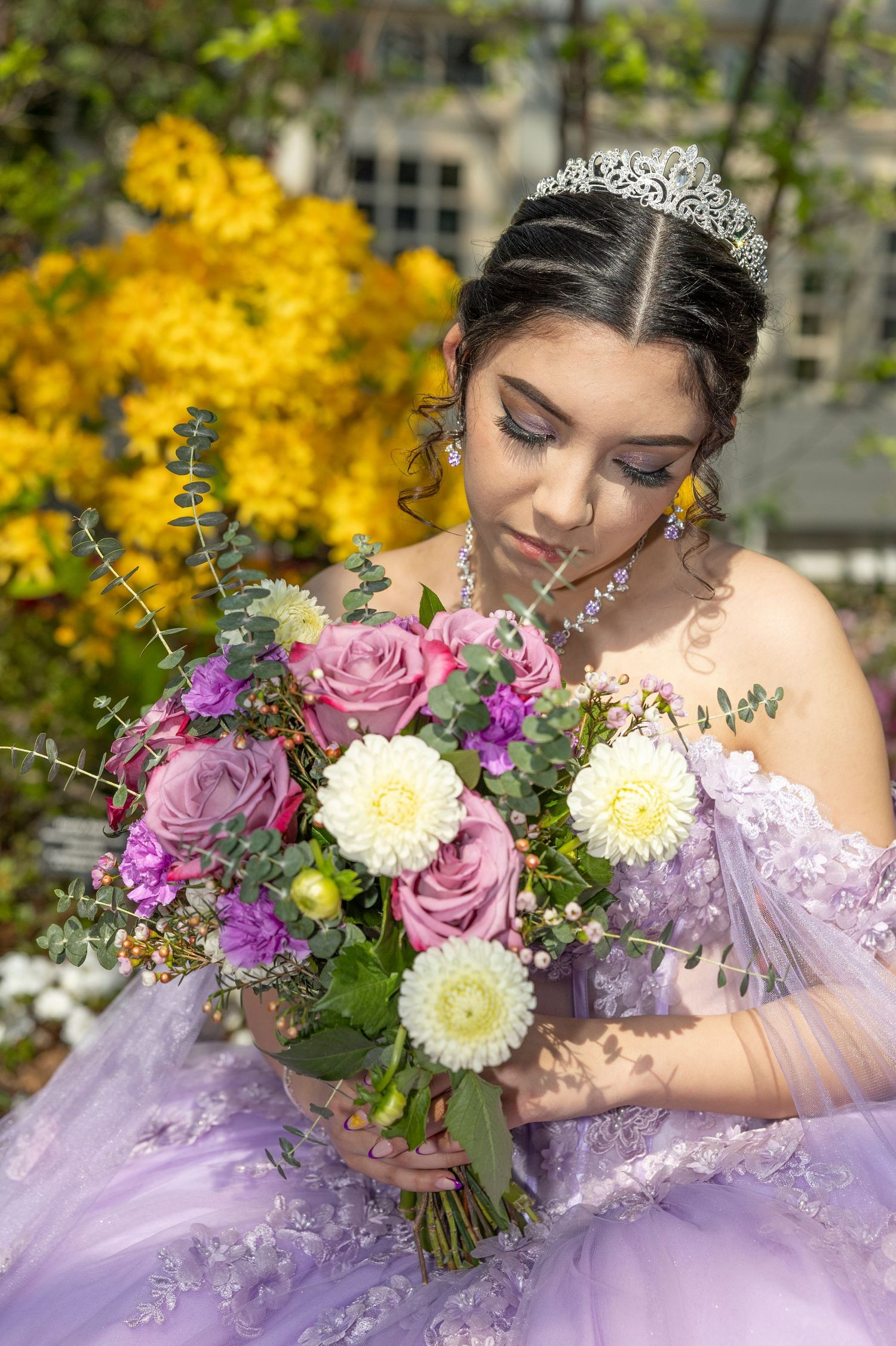 A quinceañera in a lavender ballgown poses with a pink and purple floral bouquet against a backdrop of yellow autumn foliage.