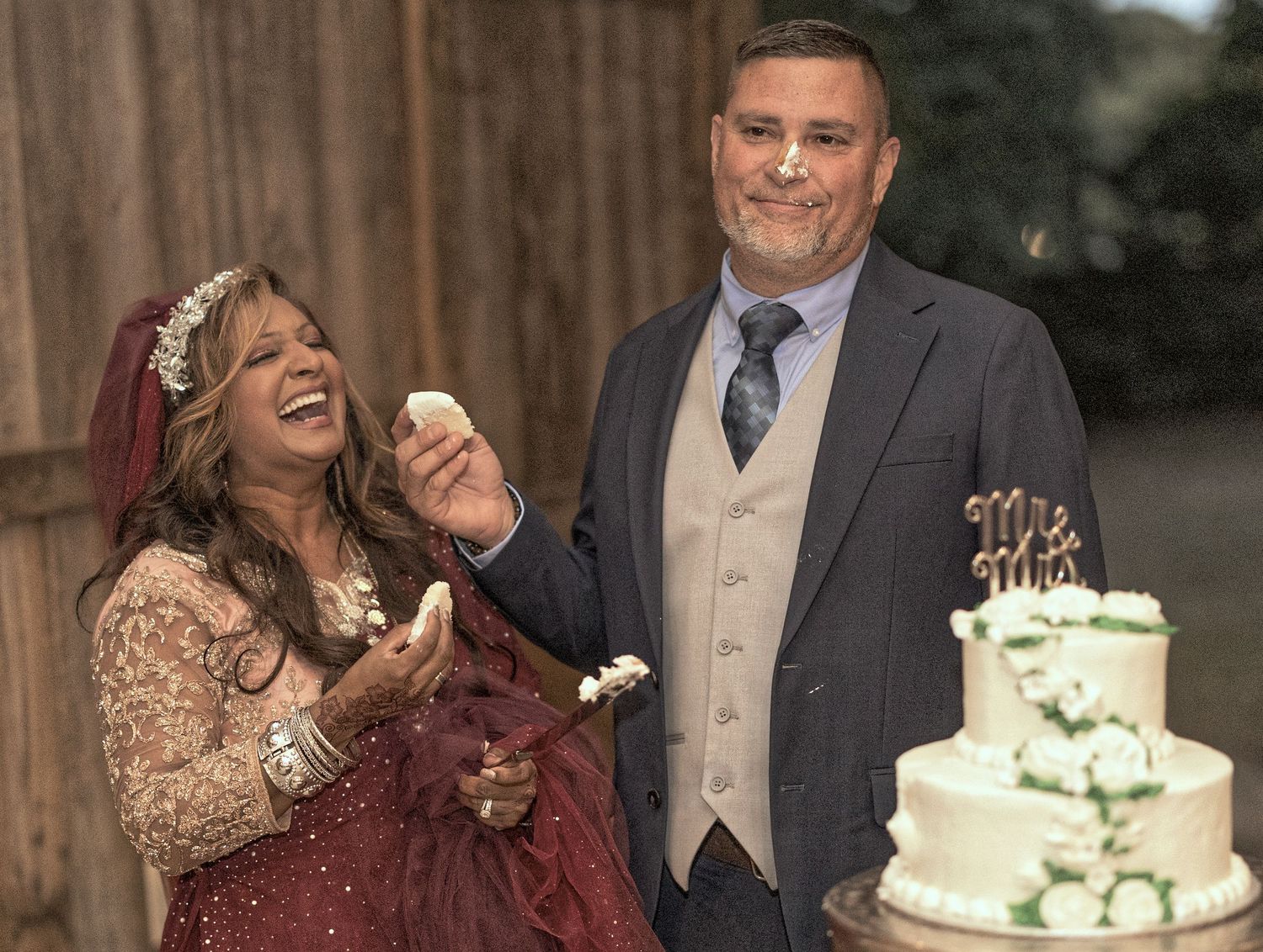 A joyful couple shares a laugh while standing next to their elegant three-tiered wedding cake at their reception.