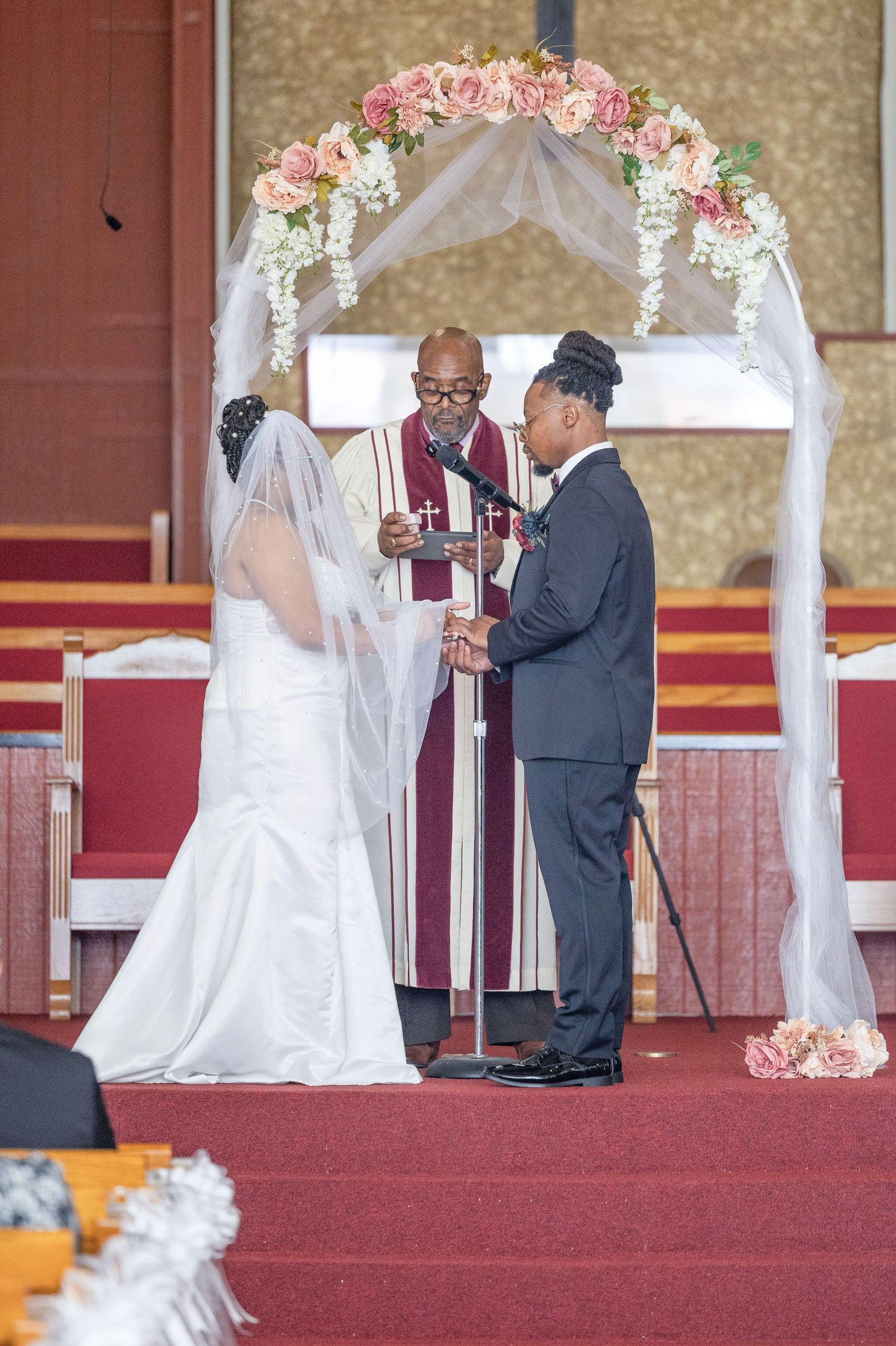 A wedding ceremony takes place under a floral arch with pink and white roses during an indoor church service.